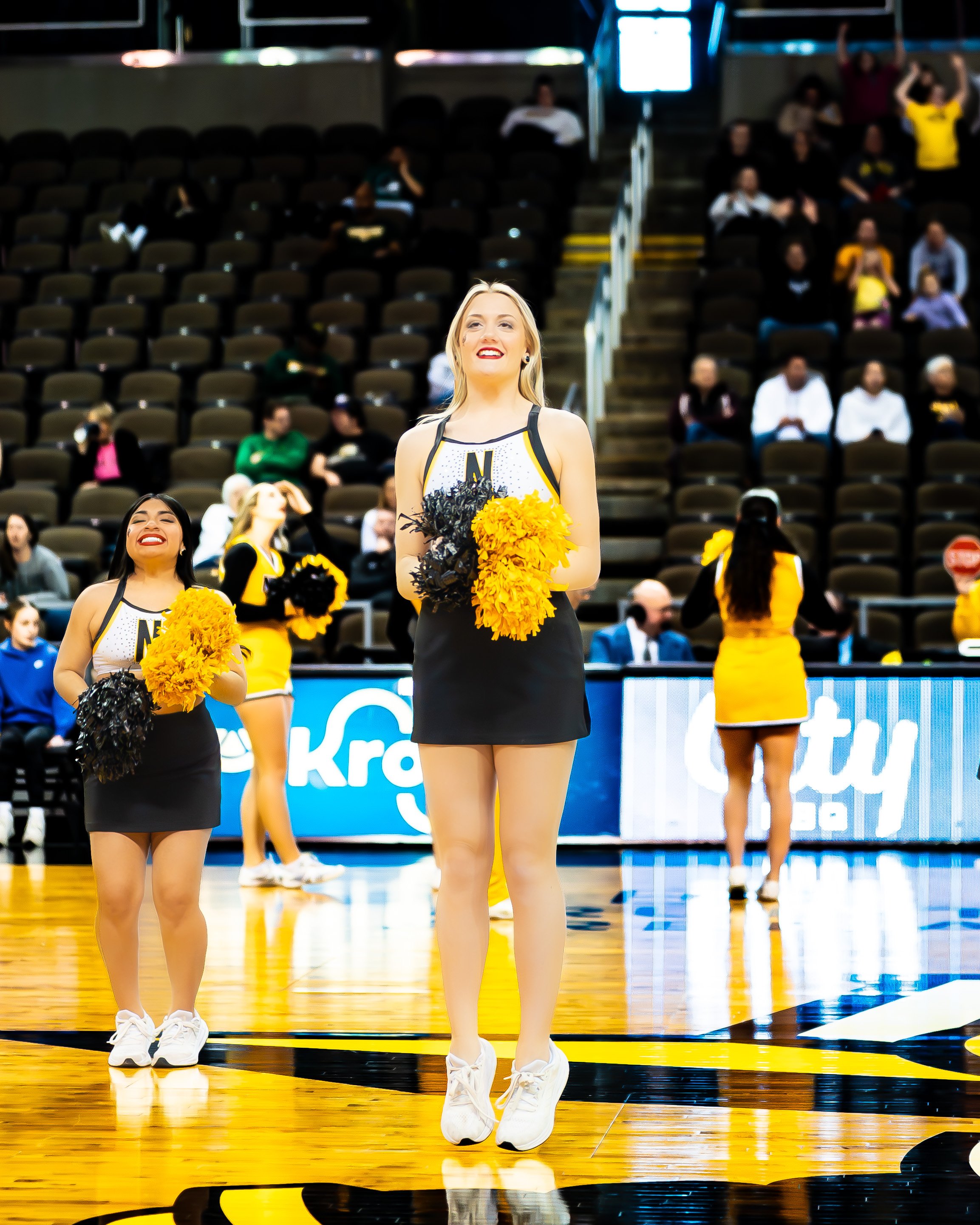 Cheerleaders performing on a basketball court during a game, holding yellow and black pom-poms, wearing black and yellow uniforms, and smiling.