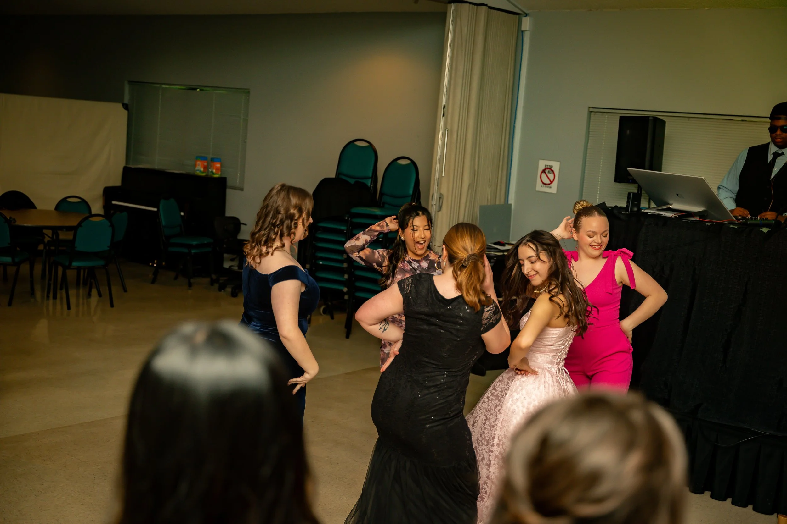 Group of women dancing and smiling at a social event in a crowded room with chairs and a DJ setup in the background.