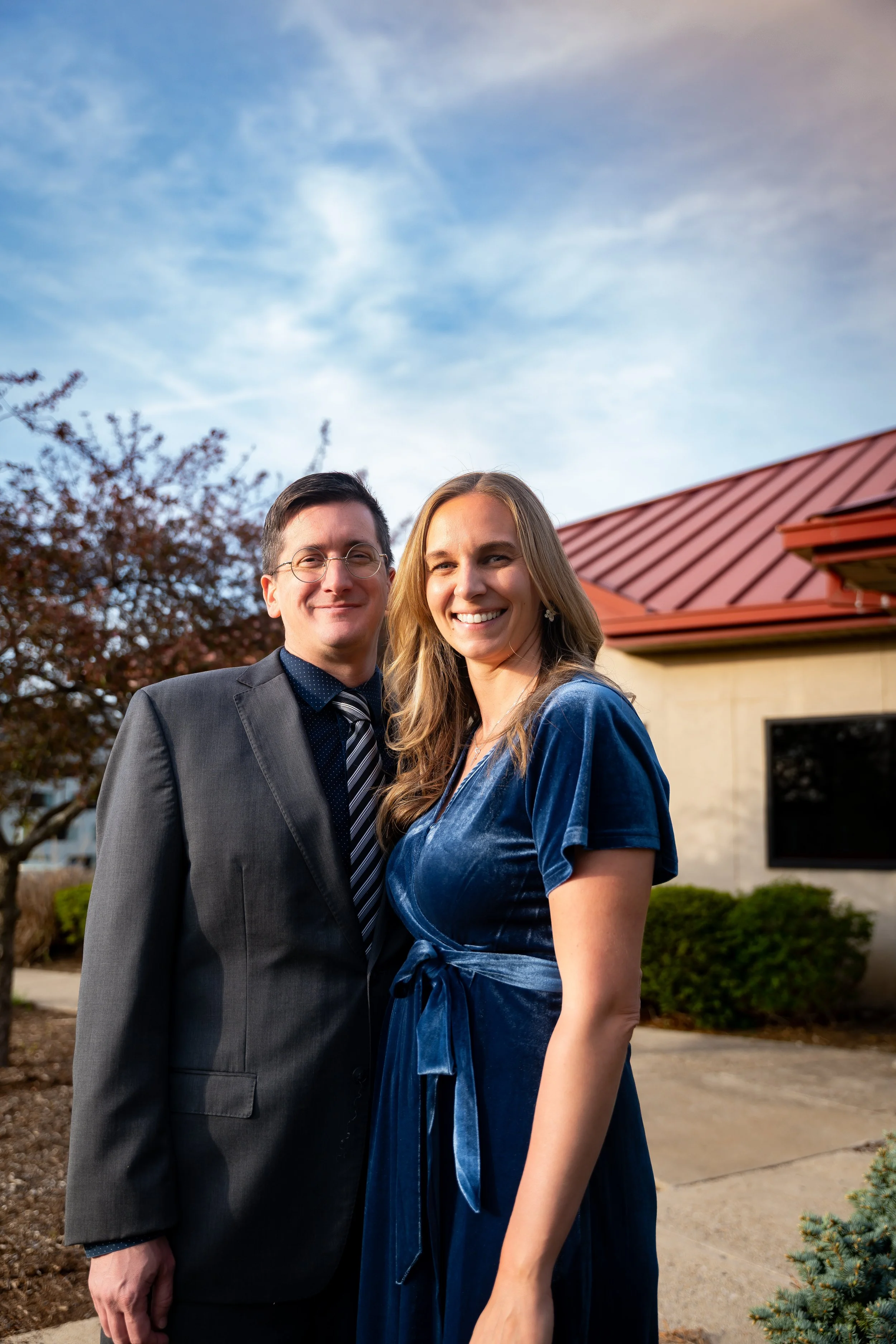 A happy man in a suit and a woman in a blue dress standing outdoors in front of a building with a red roof and trees, smiling at the camera.