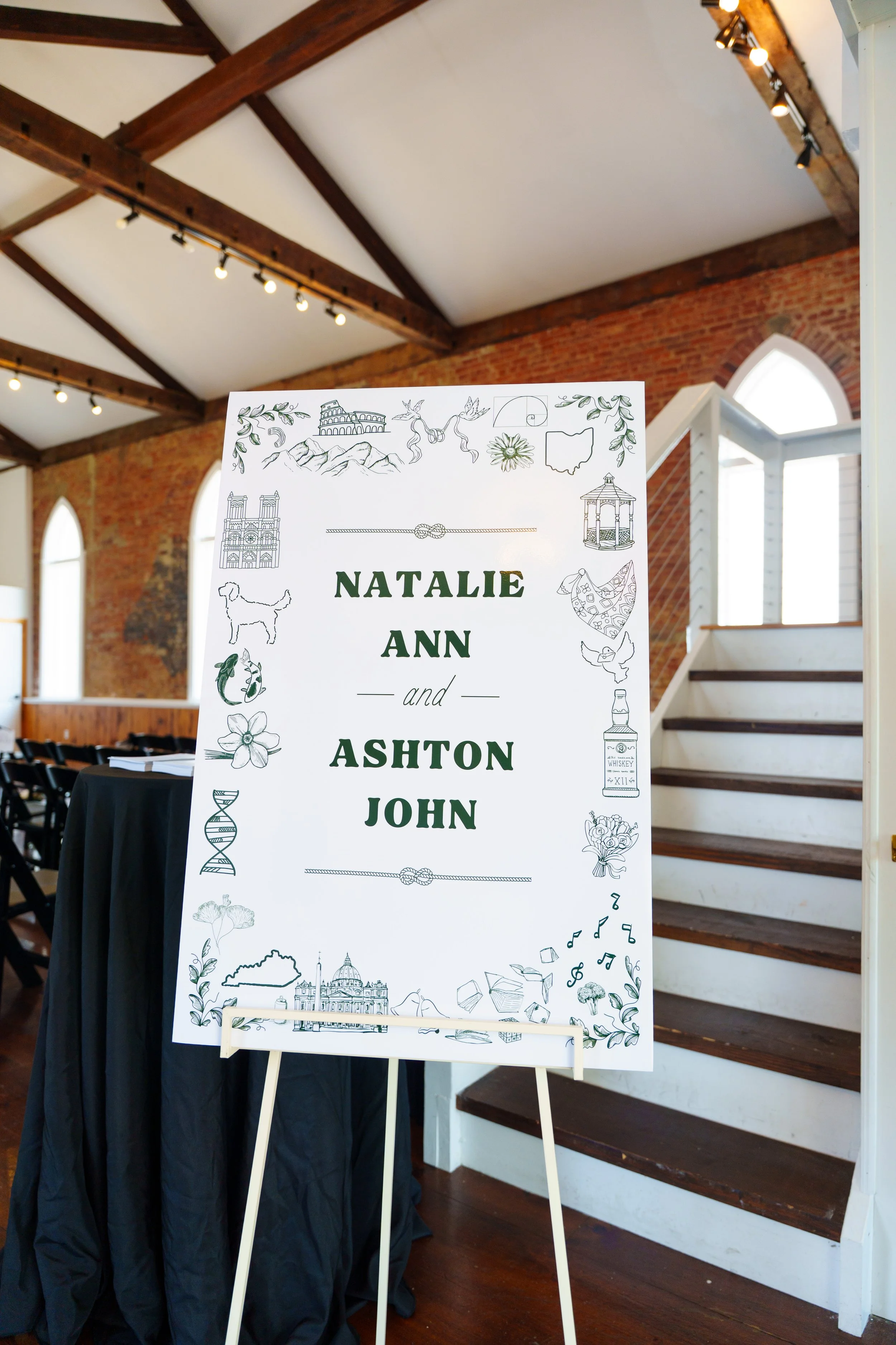 Wedding sign with the names Natalie Ann and Ashton John, decorated with illustrations of landmarks, animals, and symbols in black and white, displayed on an easel inside a venue with brick walls and arched windows.