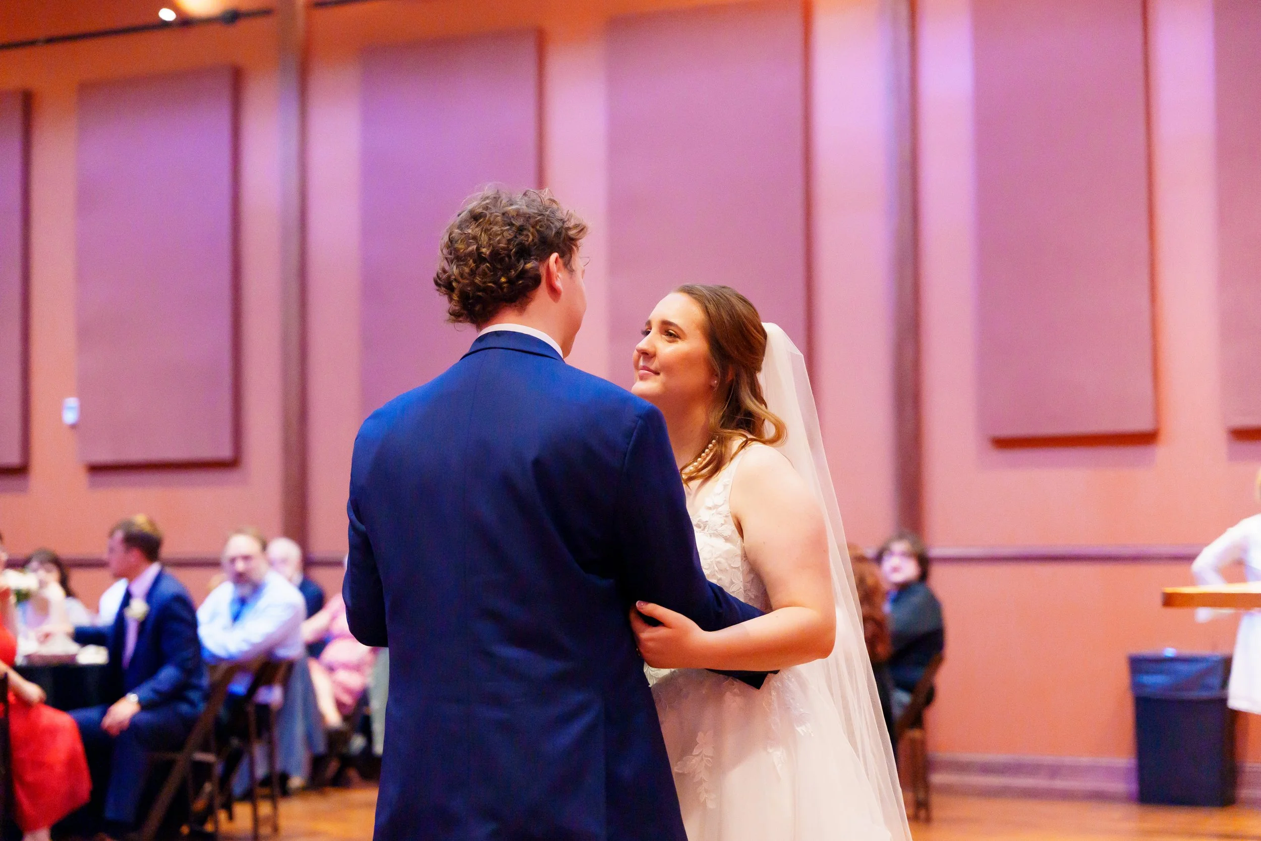 A bride and groom are holding each other and dancing at their wedding reception in a decorated hall with pink walls. There are guests seated at tables in the background.