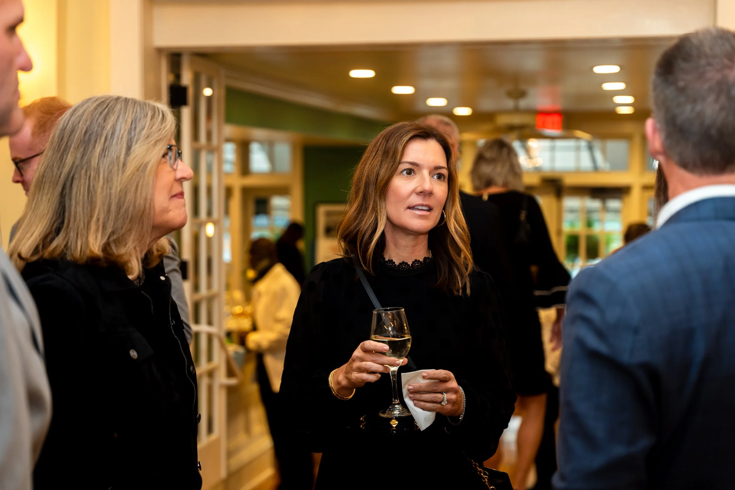 A woman with brown hair holding a glass of white wine engaged in conversation at a social event, with other people in the background.