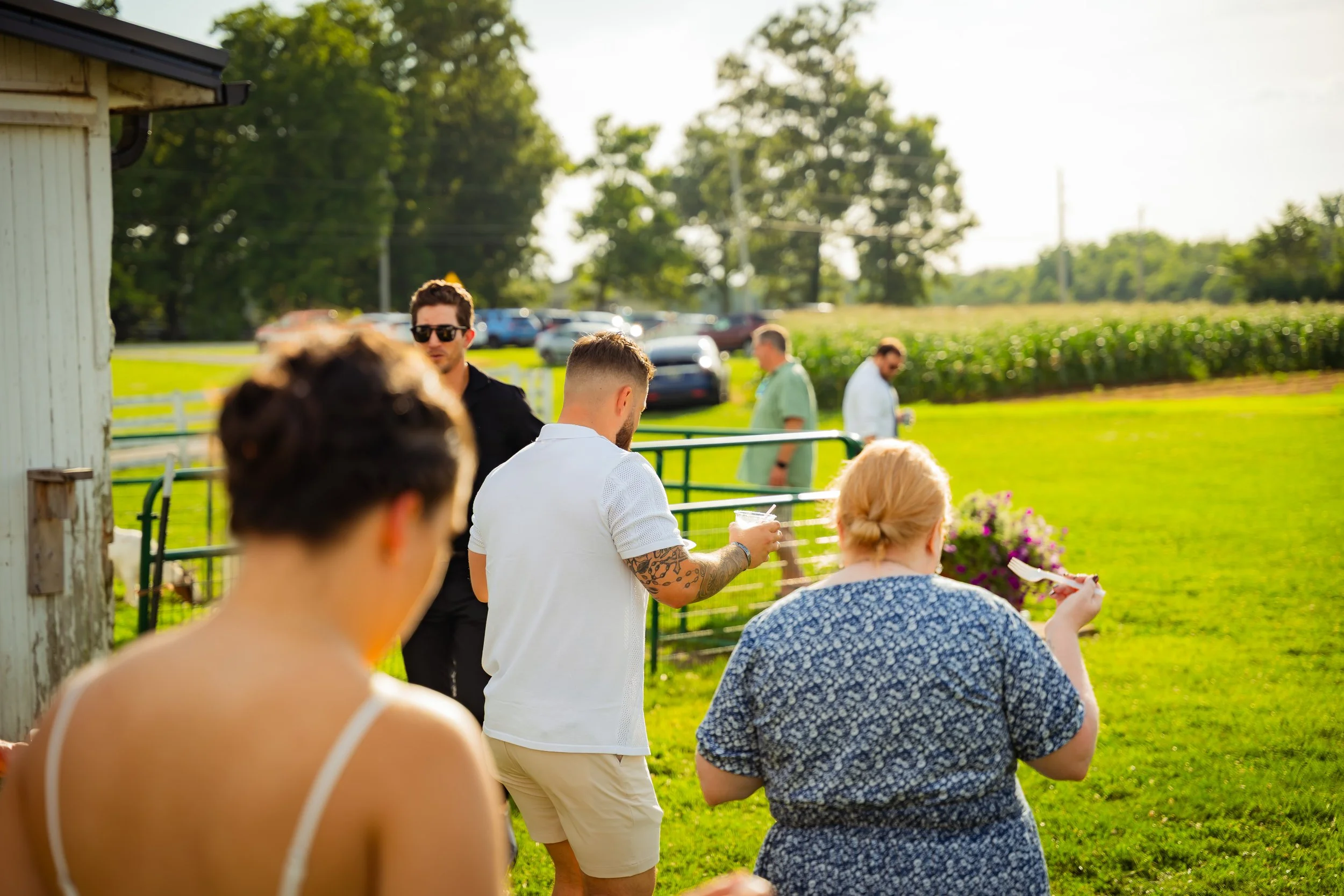 People gathered outdoors near a small white building, some sitting and some standing, with a field and trees in the background. The scene appears to be a casual social event on a sunny day.