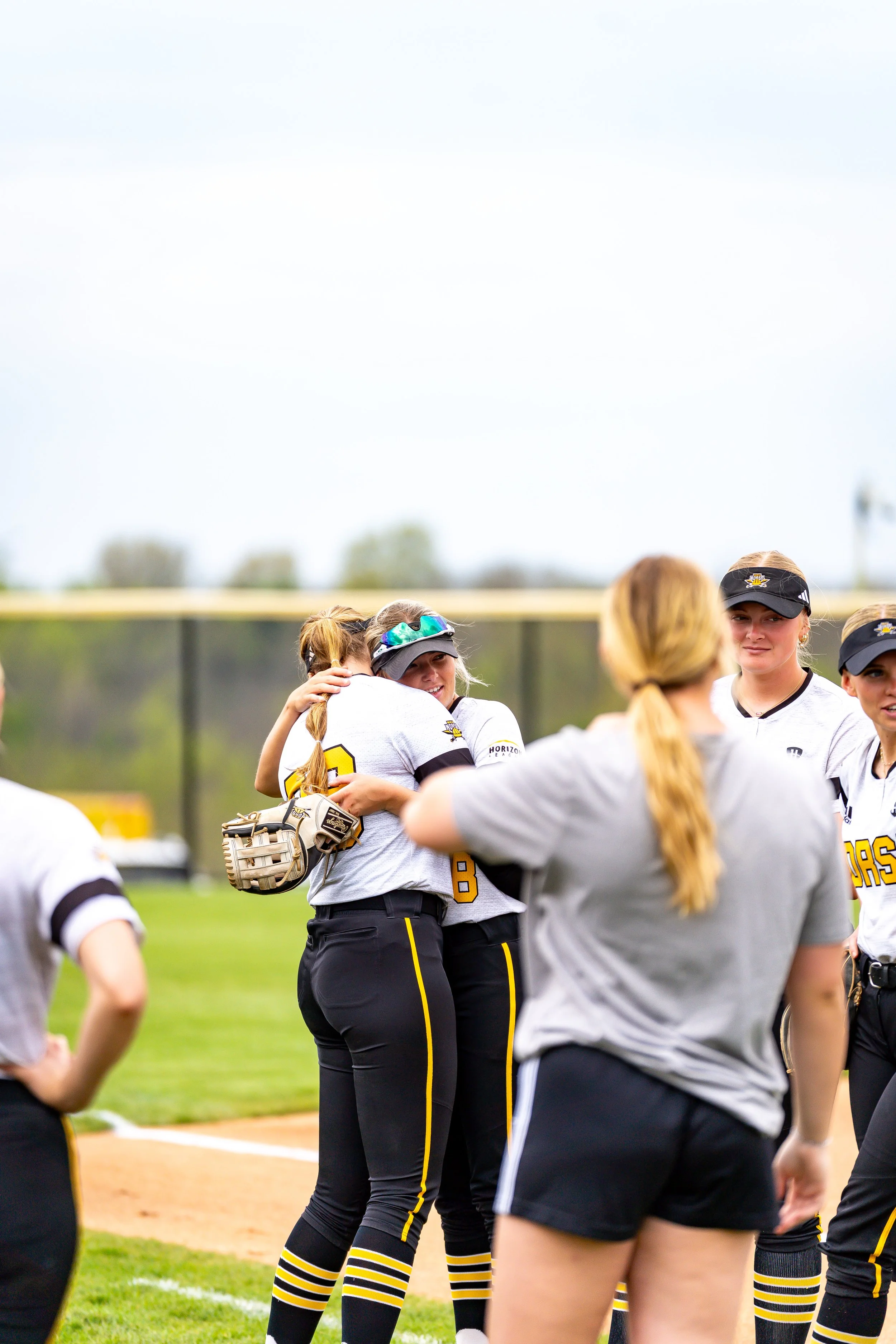 Women softball players in black and yellow uniforms standing on a baseball field during a game, with a player hugging another.