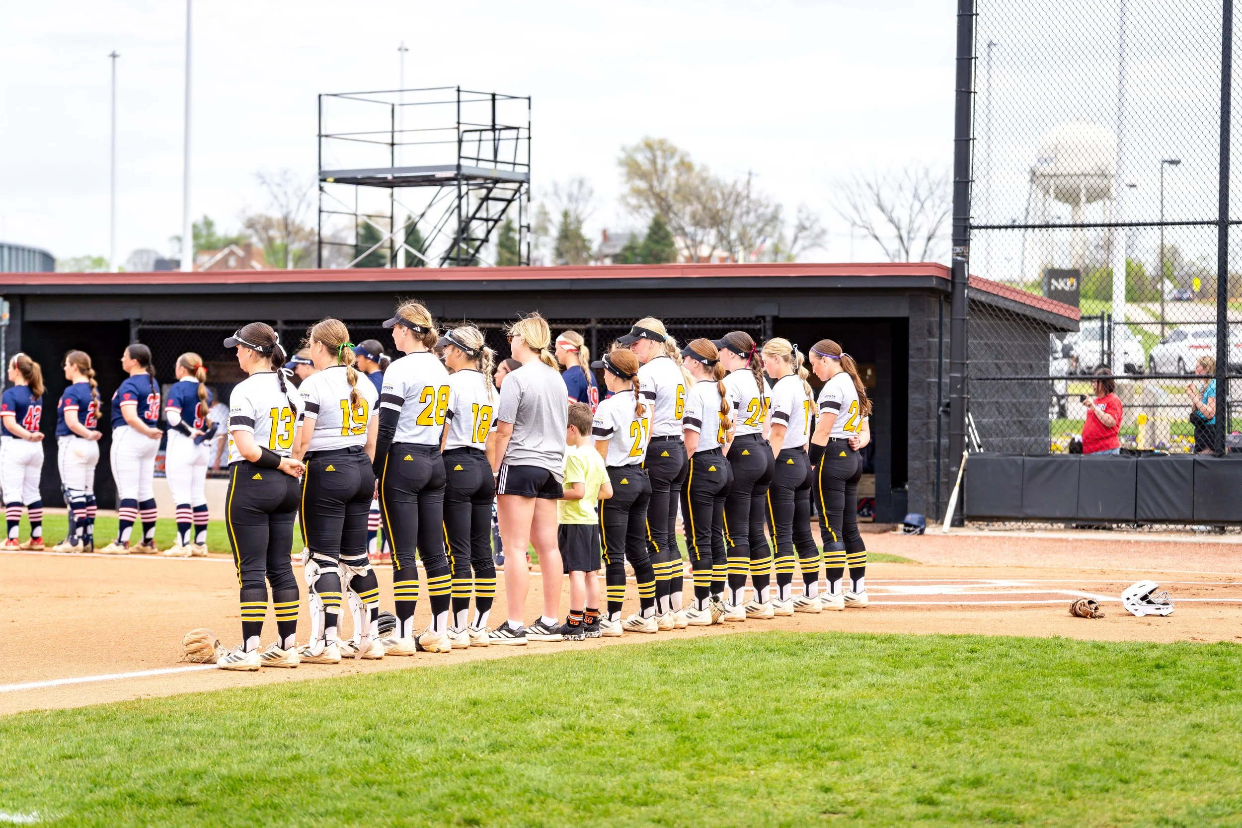 Girls' softball team standing in line on the field, some in uniforms with numbers and stripes, and some wearing gray shirts with black shorts, at a baseball field with a black dugout, fence, and trees in the background.