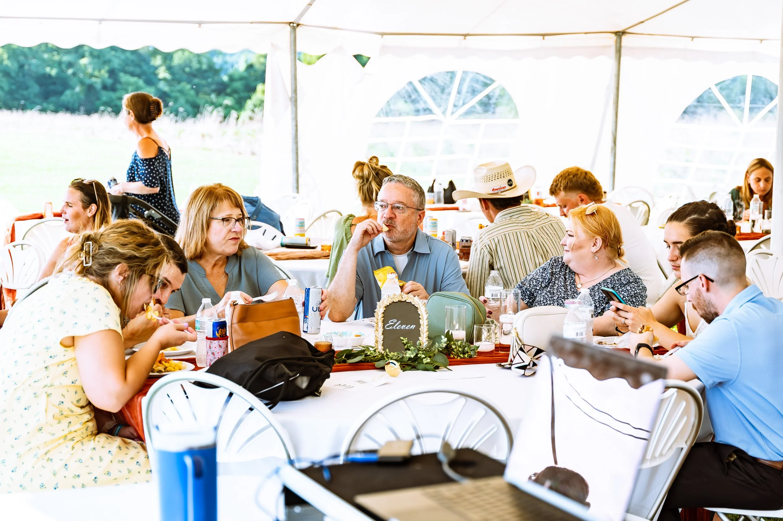 People sitting at a table inside a large white tent outdoors, enjoying a meal and conversation on a sunny day.
