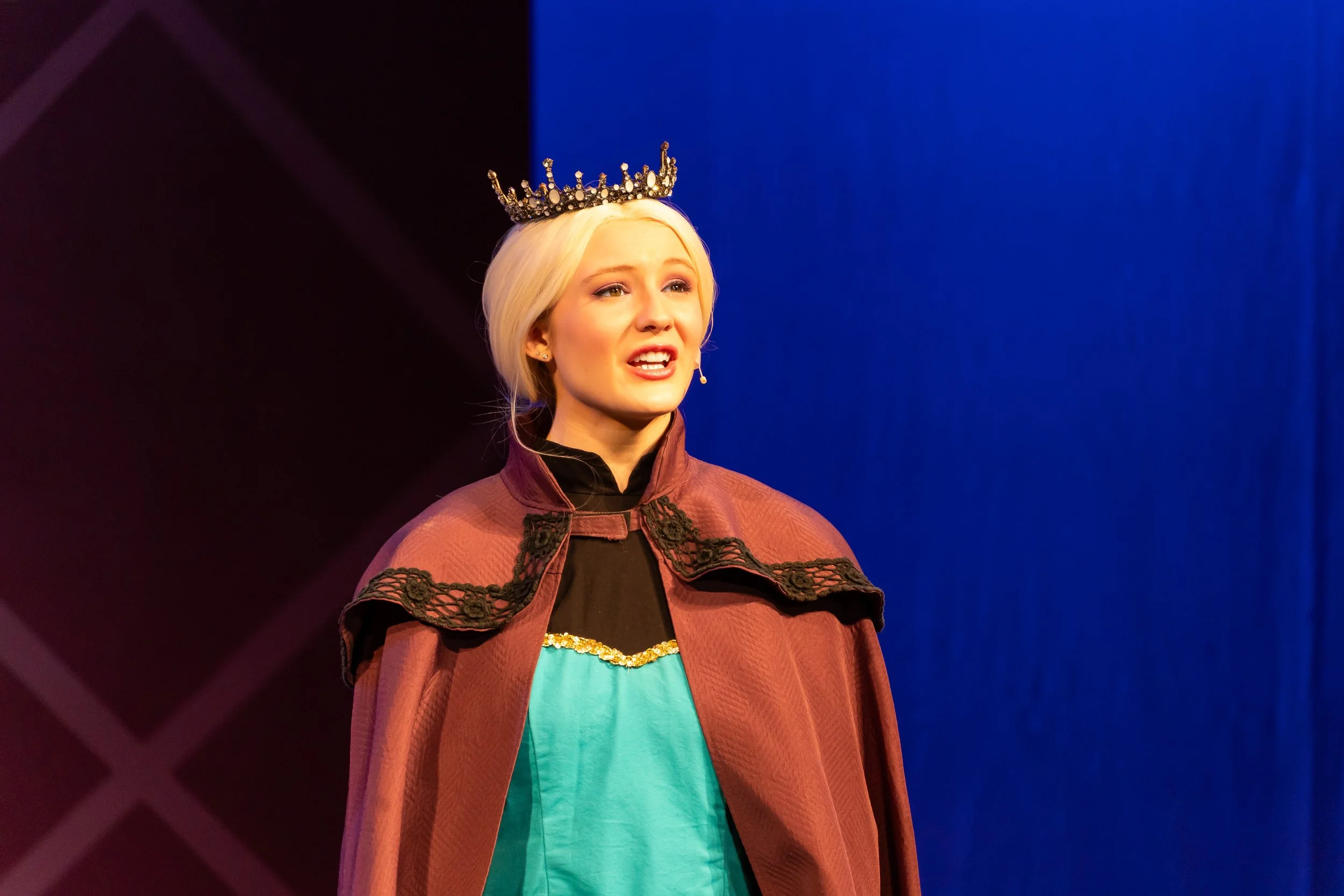 A woman with platinum blonde hair wears a crown and speaking into a microphone, dressed in a red coat with black lace details and a teal top, standing on a stage with a blue background and dark area to her left.