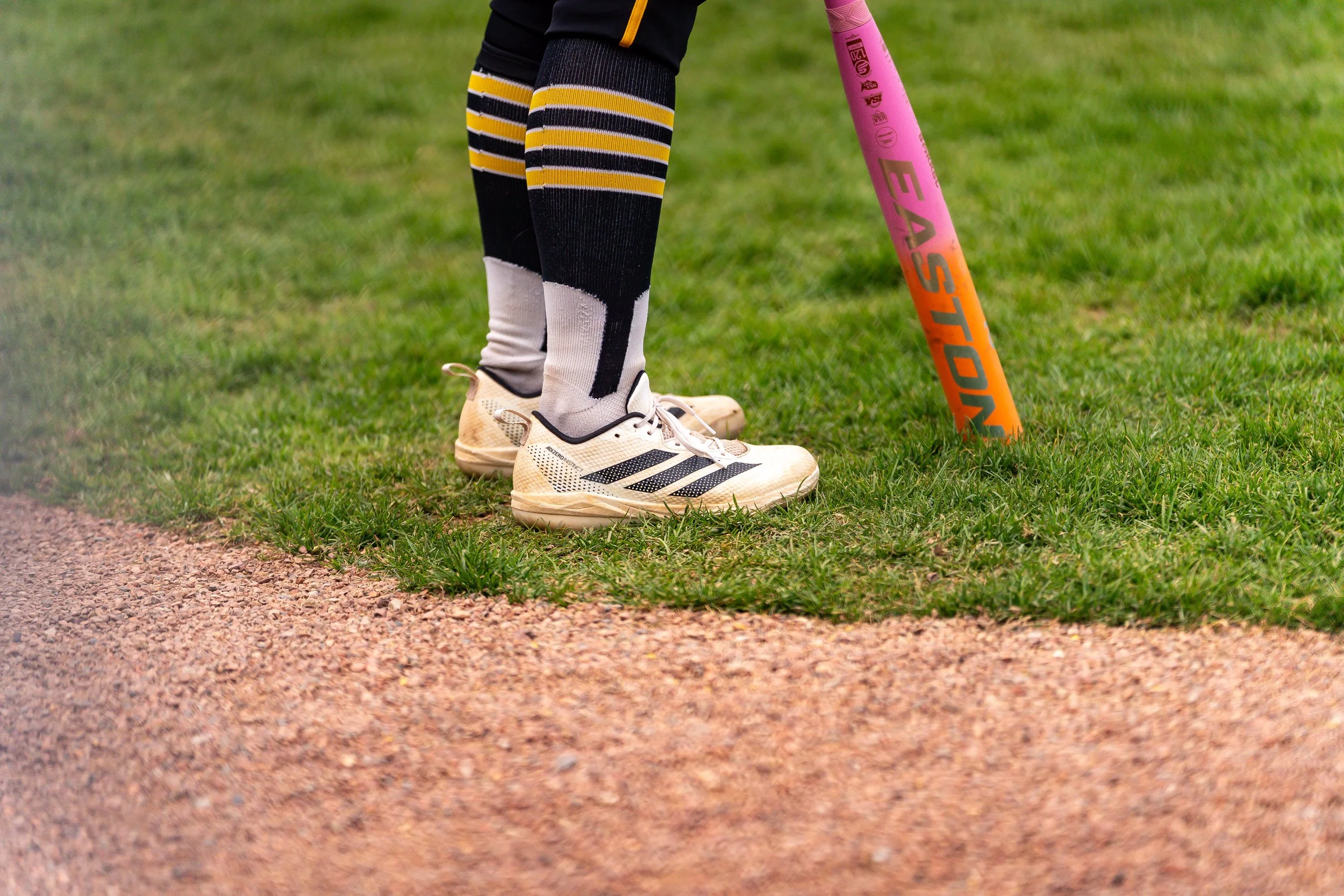 Close-up of a person’s legs wearing black, yellow, and white sports socks and beige athletic shoes, standing on a grassy field beside a gravel path, holding a pink and orange field hockey stick.