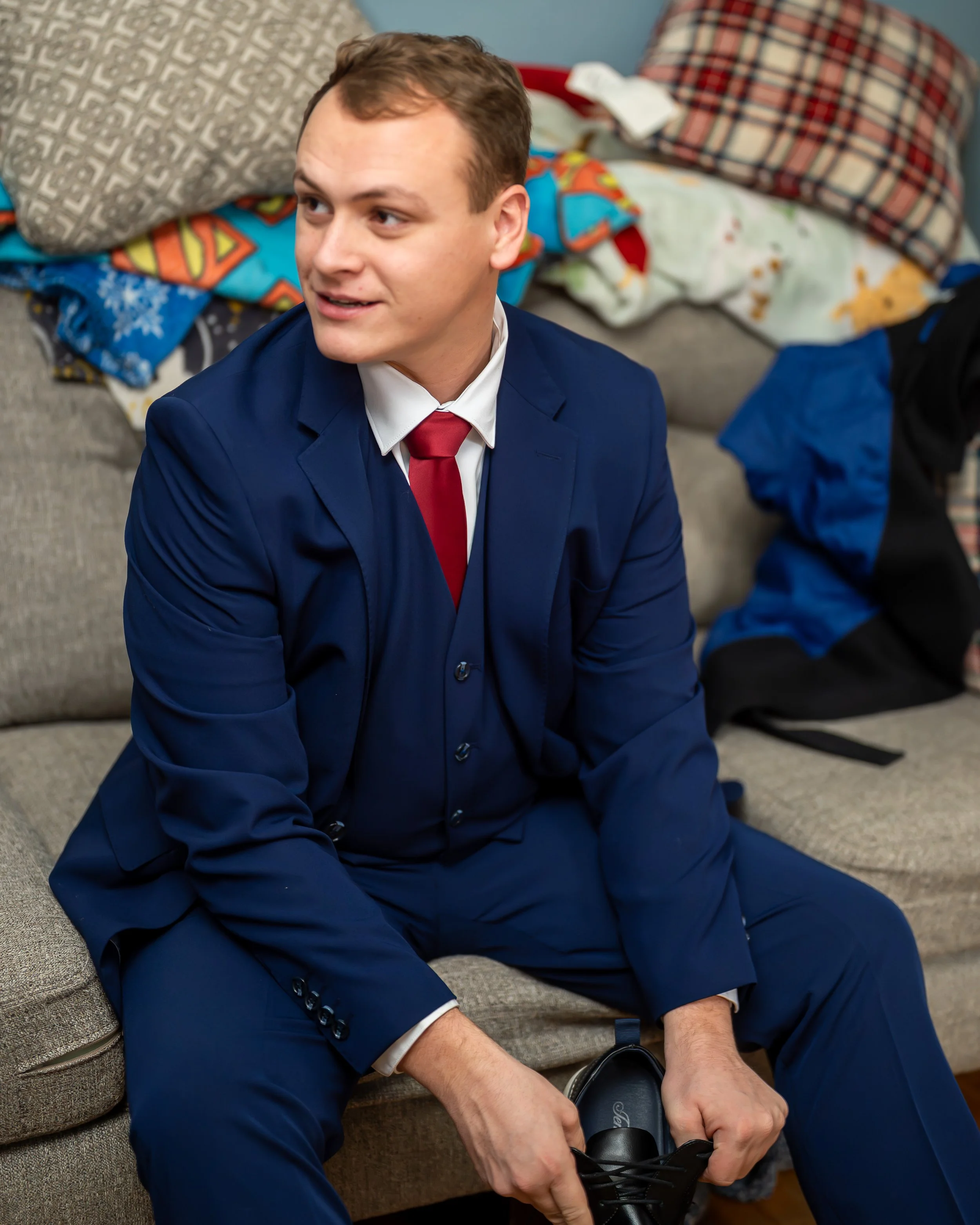 Young man in a blue suit tying his shoelaces while sitting on a gray couch. The background has piles of laundry and a blue wall.