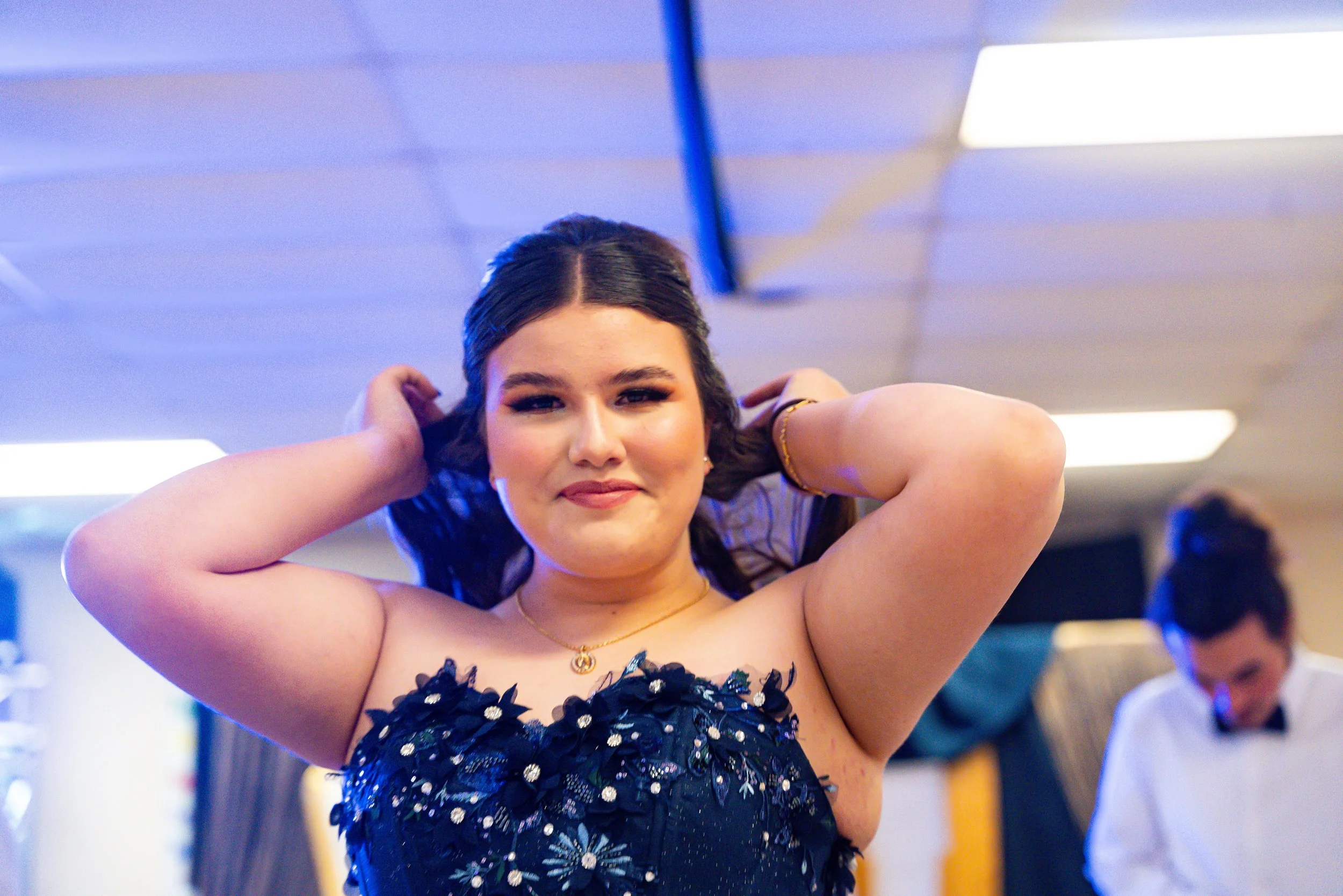 A young woman with dark hair and makeup, wearing a black dress with floral embellishments, smiling and posing with her arms behind her head in an indoor setting with colorful lighting and a blurred background.
