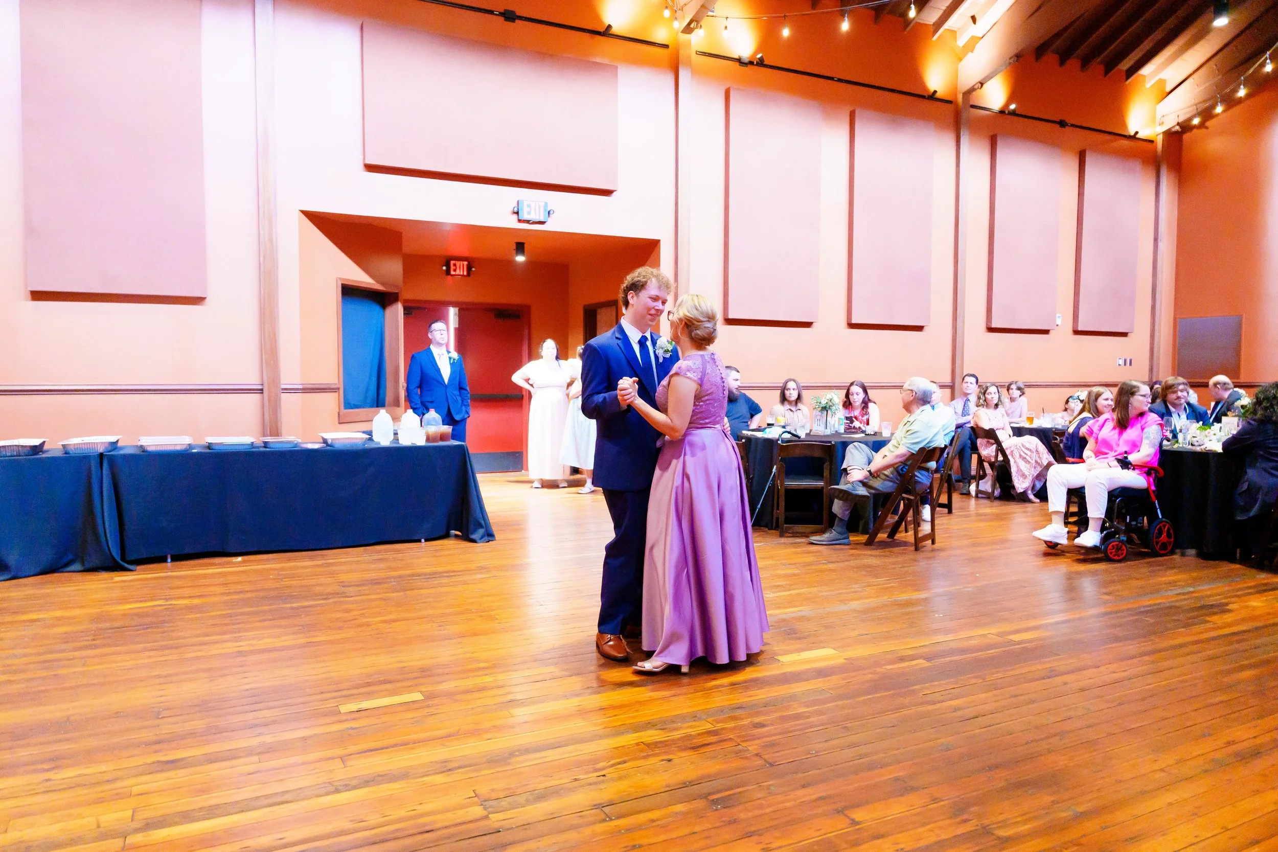 A couple dancing at a wedding reception in a large, brightly colored room with high ceilings and string lights. Guests are seated at tables watching them.