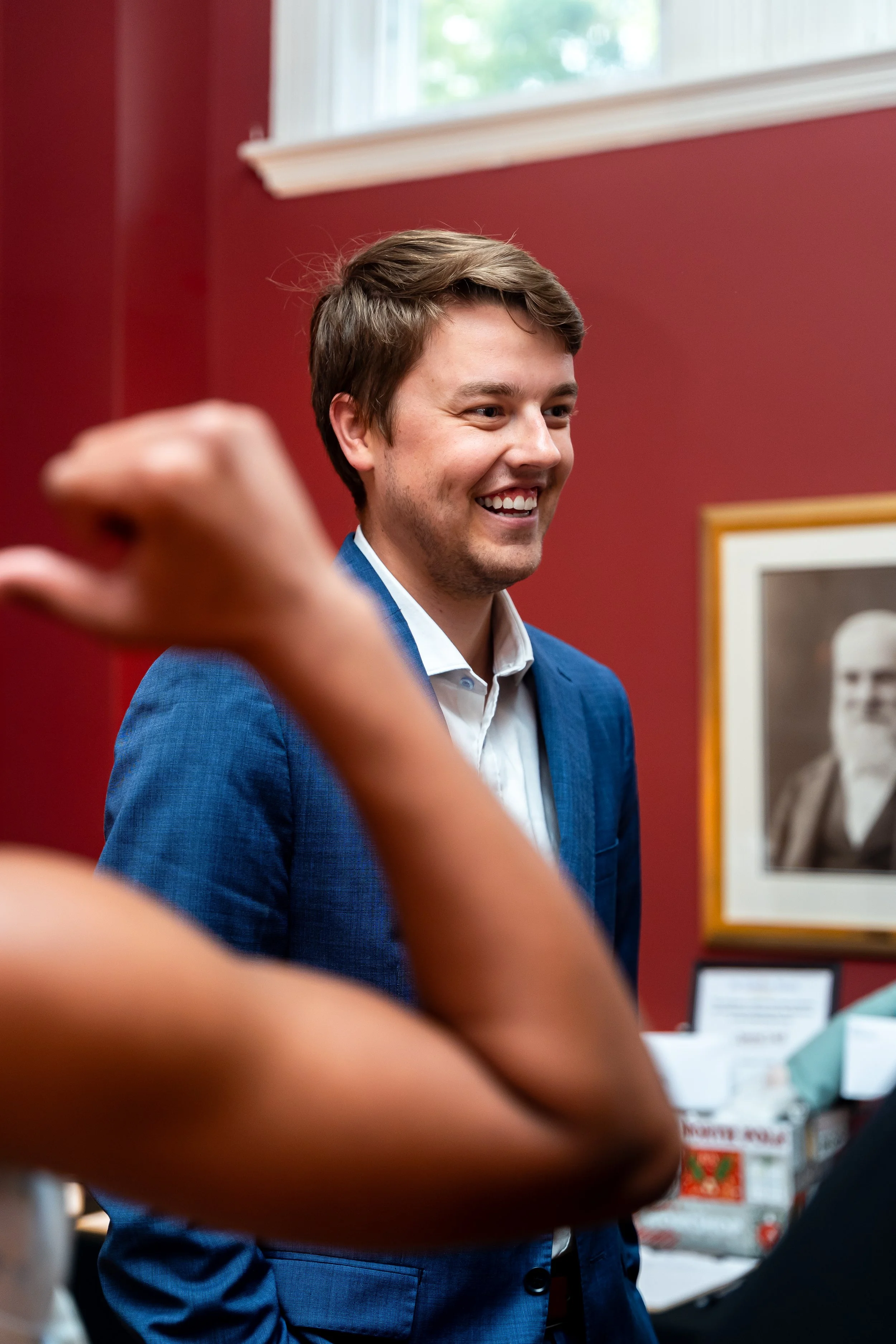 A young man with brown hair, smiling, wearing a blue blazer and white shirt, standing indoors with a red wall and window behind him.
