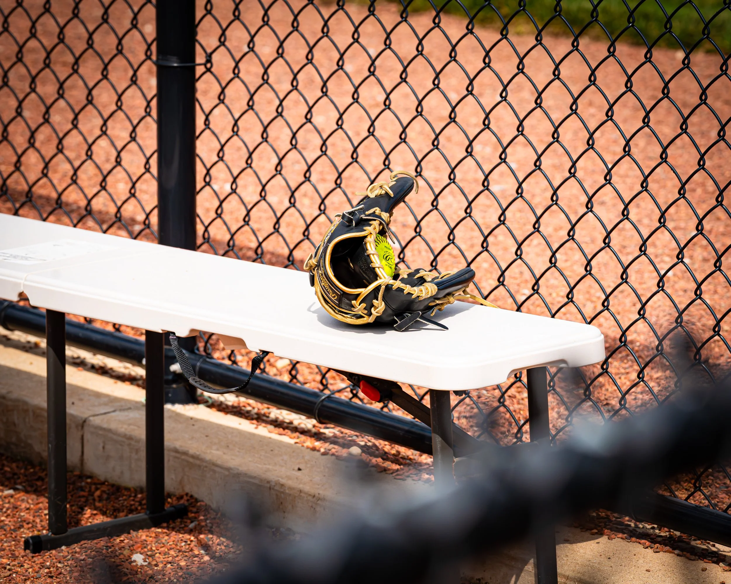 A black baseball glove resting on a white bench outdoors with a chain-link fence and reddish dirt ground in the background.