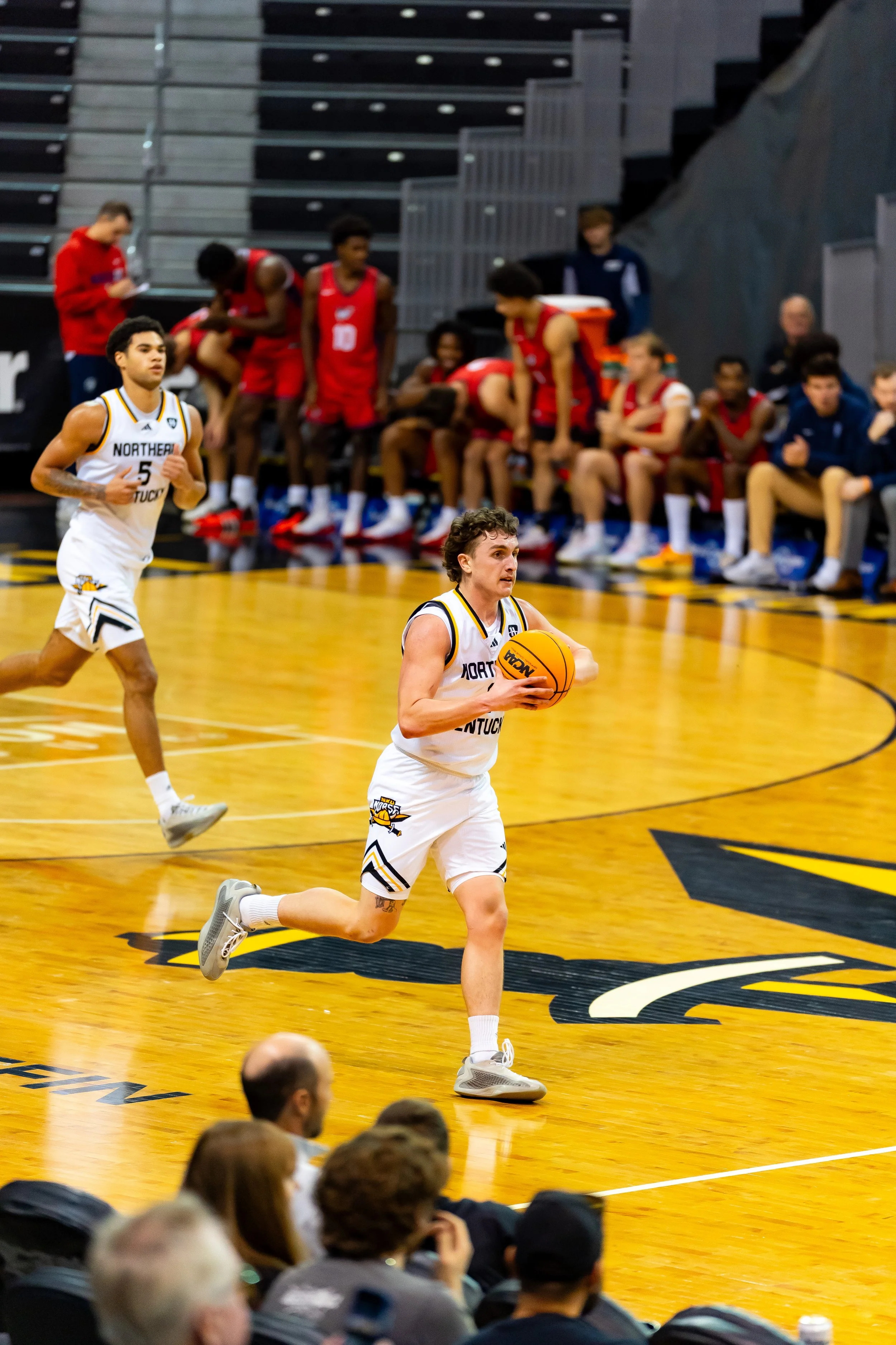 A basketball player in a white uniform running on the court while holding a basketball, with players on the bench and spectators in the background.
