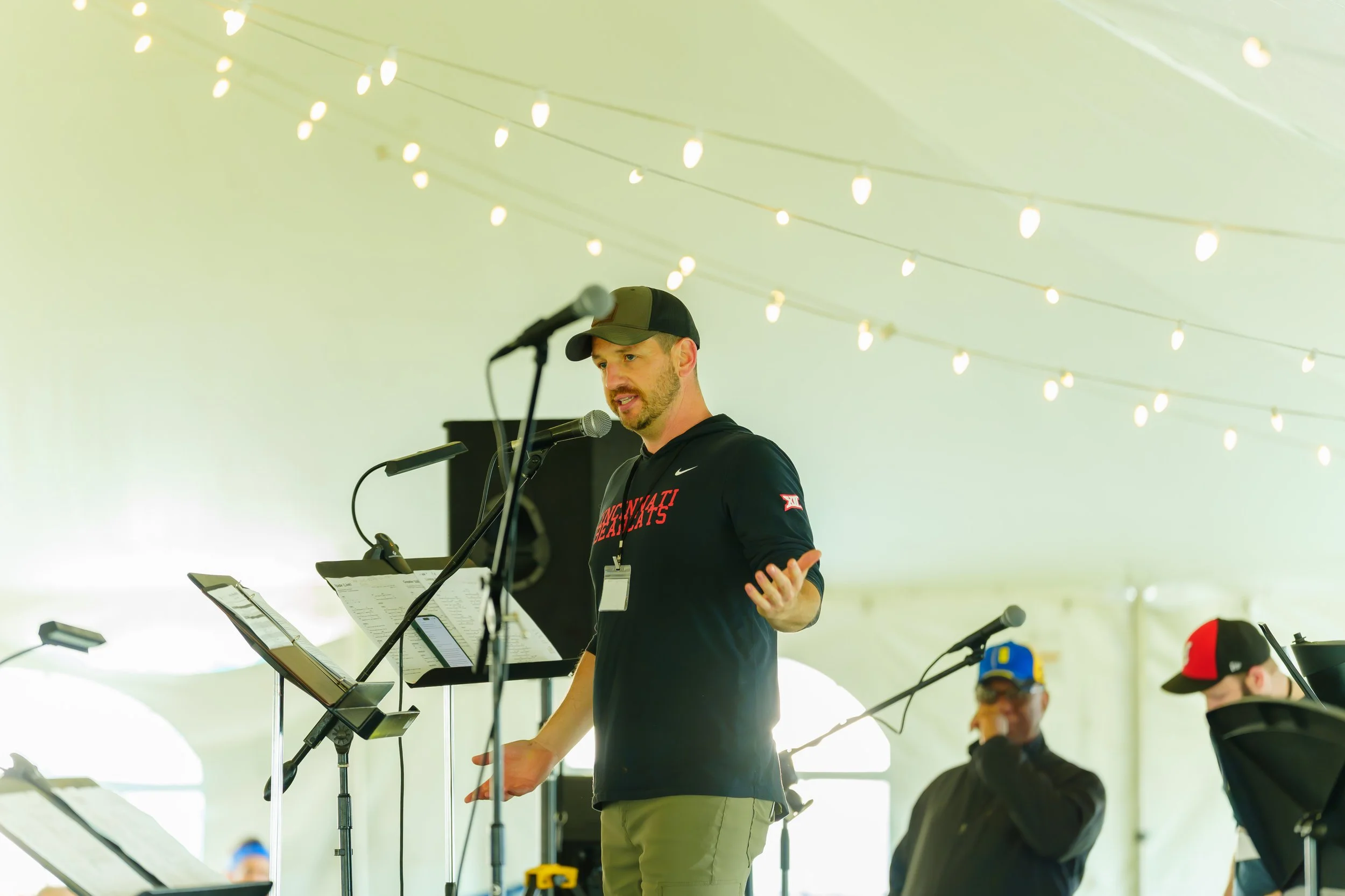 A man wearing a black cap and black hoodie speaking into a microphone at a standing music or speech event, with sheet music on music stands and string lights hanging overhead under a white tent.