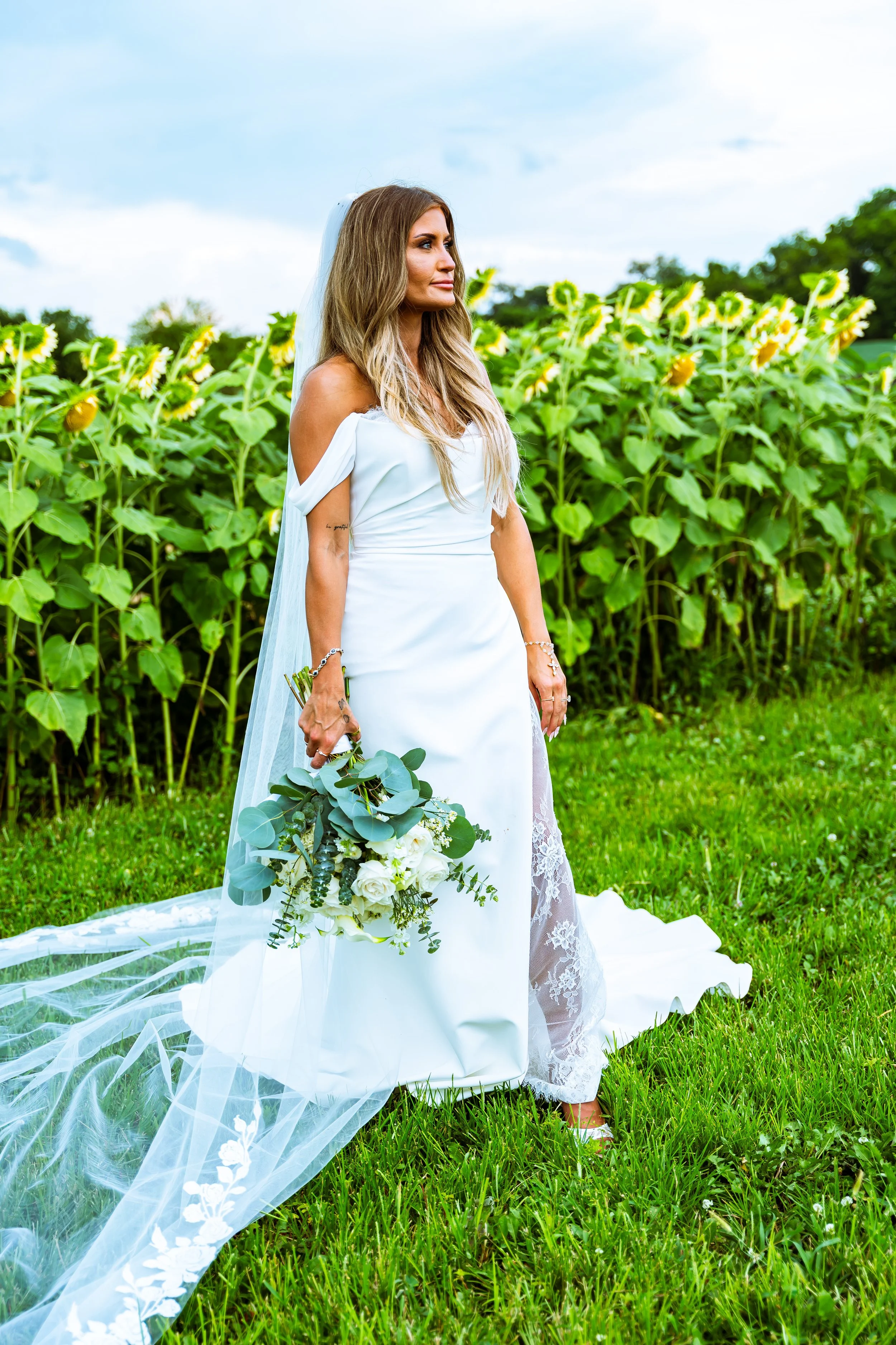 A bride standing outdoors in a white wedding dress holding a bouquet of white roses and greenery, with a sunflower field and trees in the background.