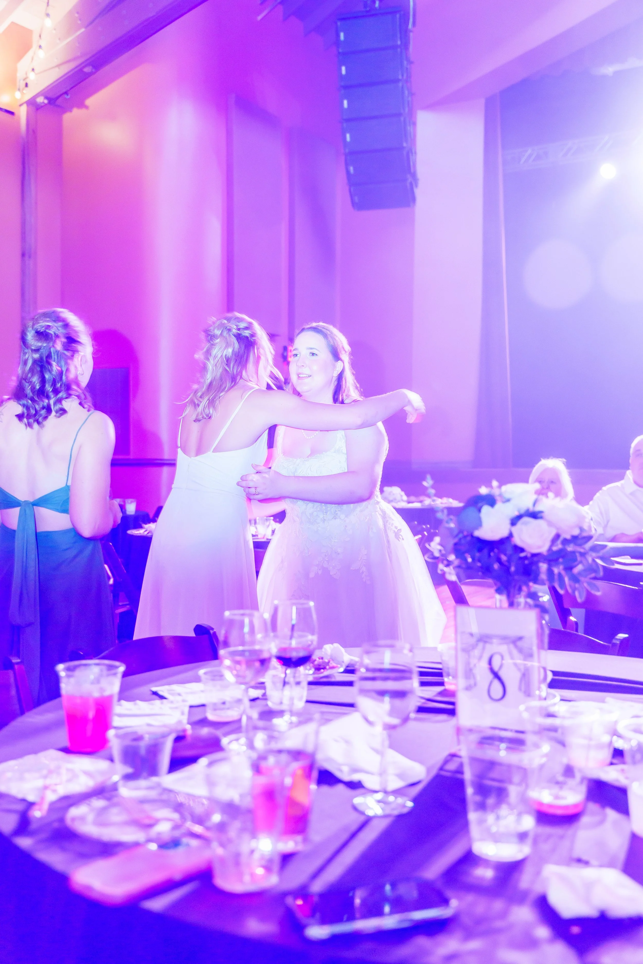 Two women dancing and chatting at a reception or party with tables set with drinks and floral centerpieces, under purple lighting.