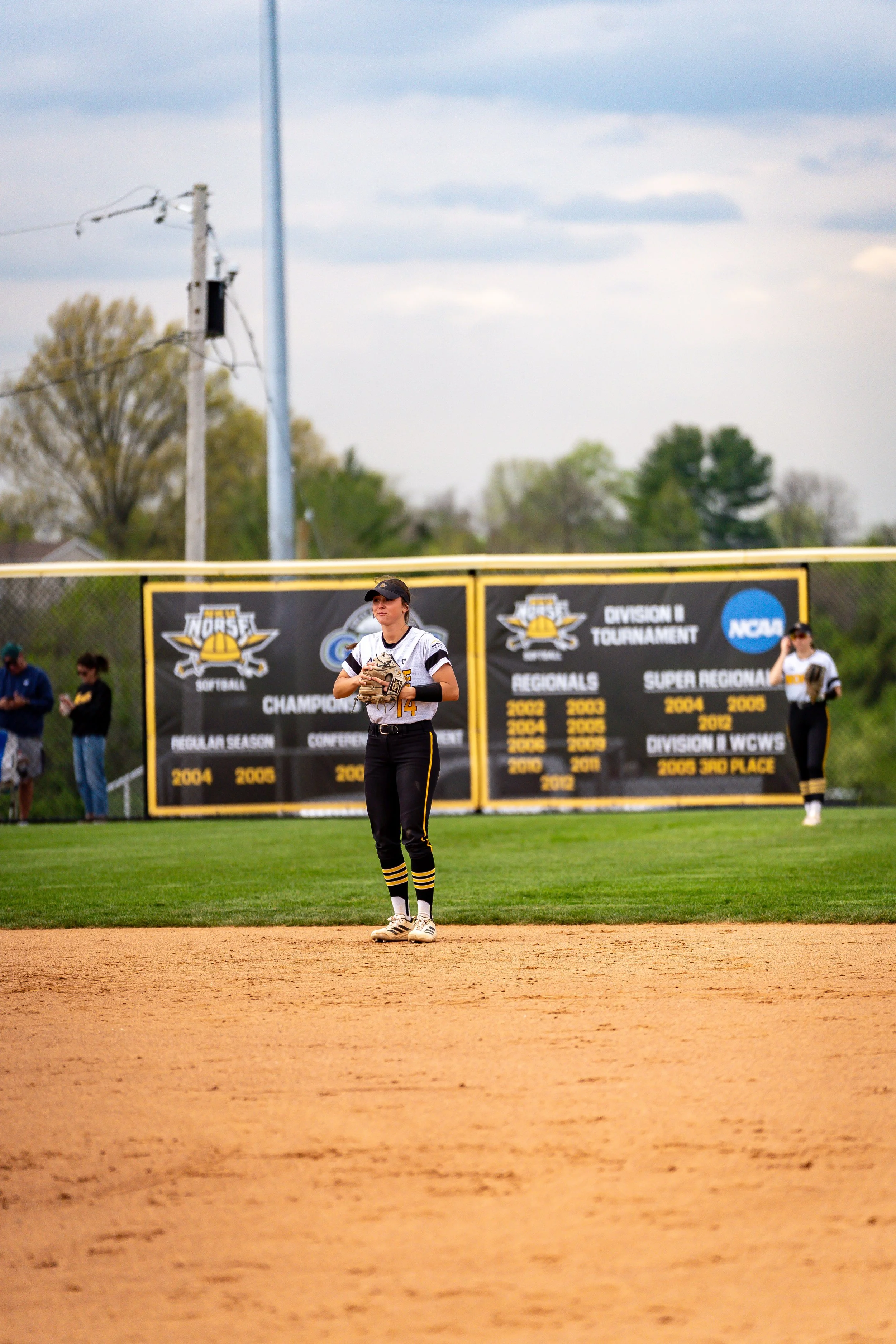 A female softball player standing on a dirt infield near the outfield fence, holding a glove and wearing a black cap, jersey, and black pants with yellow stripes, at a softball field with banners and trees in the background.