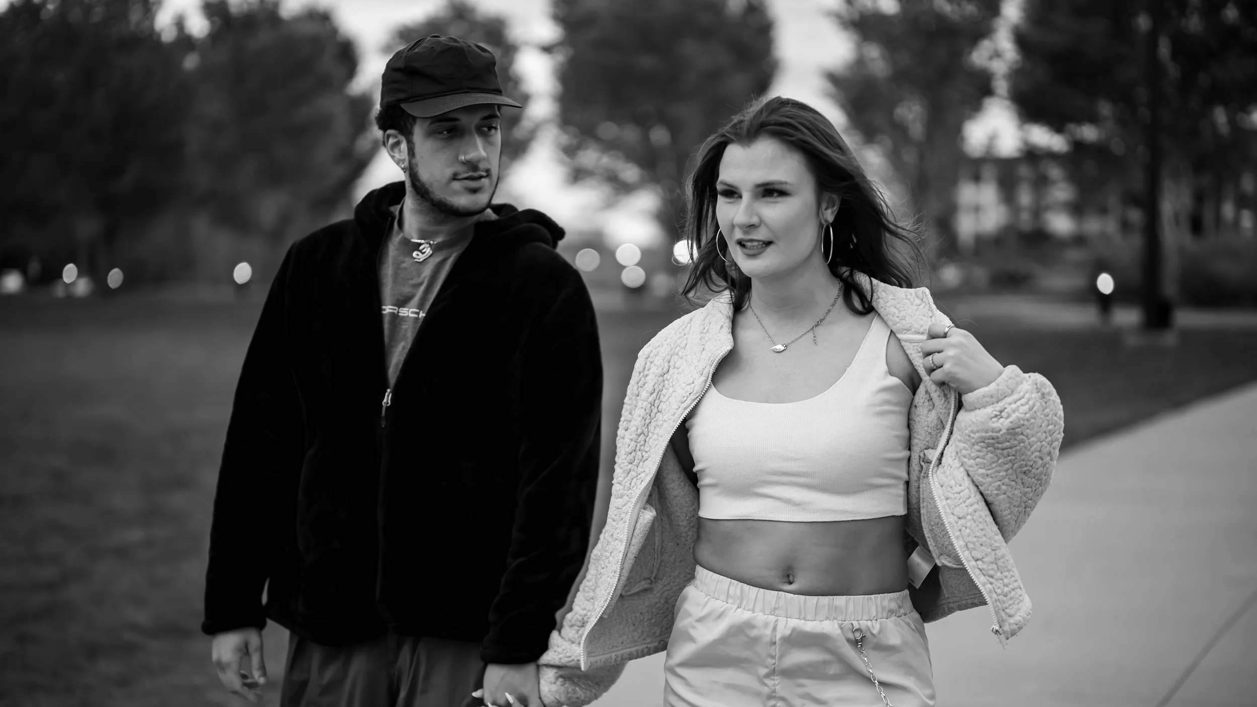 A black and white photo of a young man with a beard, wearing a cap, and a young woman with long hair, wearing hoop earrings and a crop top, walking outdoors in a park with trees in the background.