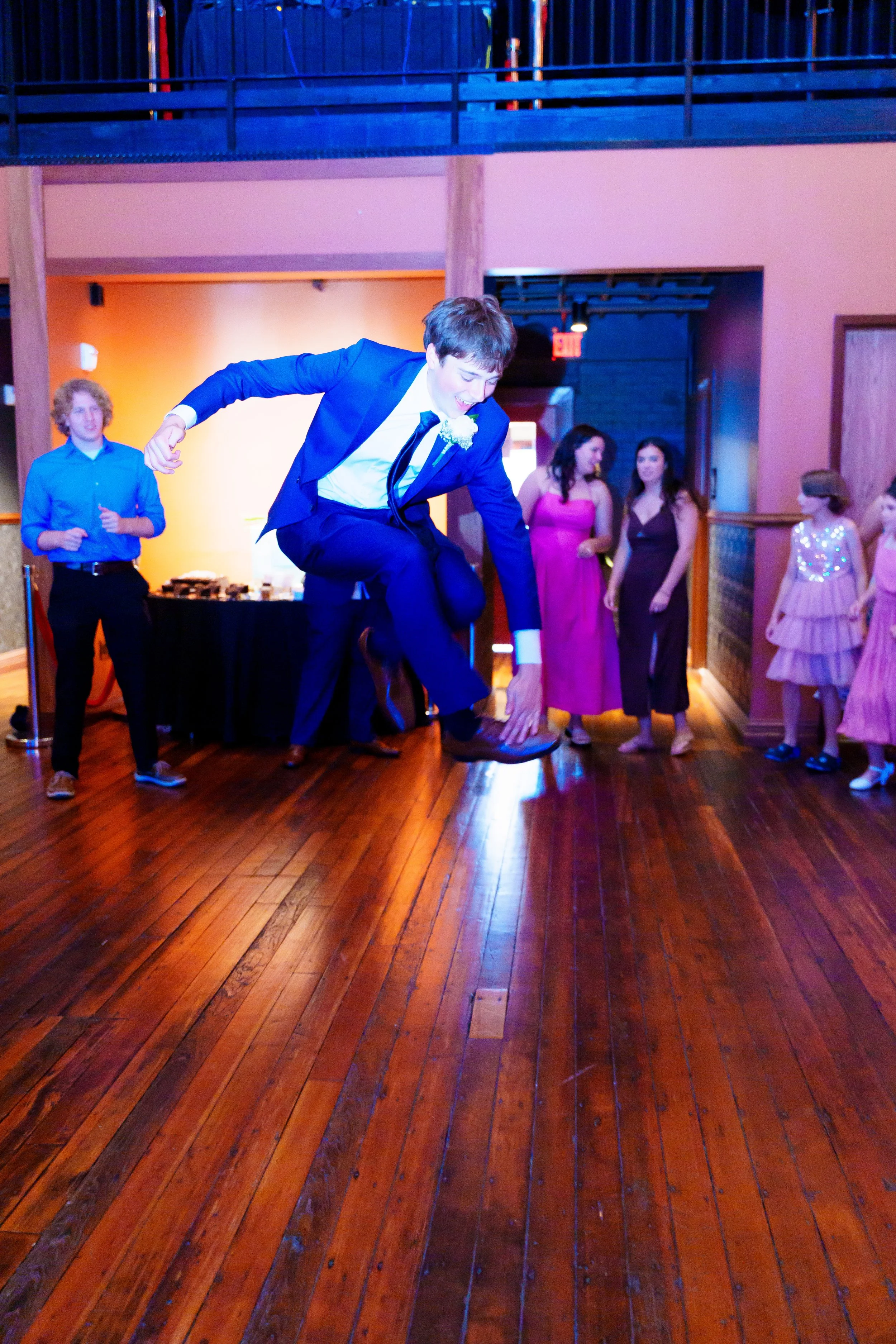 Young man in a blue suit is jumping and dancing on a wooden floor at a formal event, with several women in dresses watching in the background.