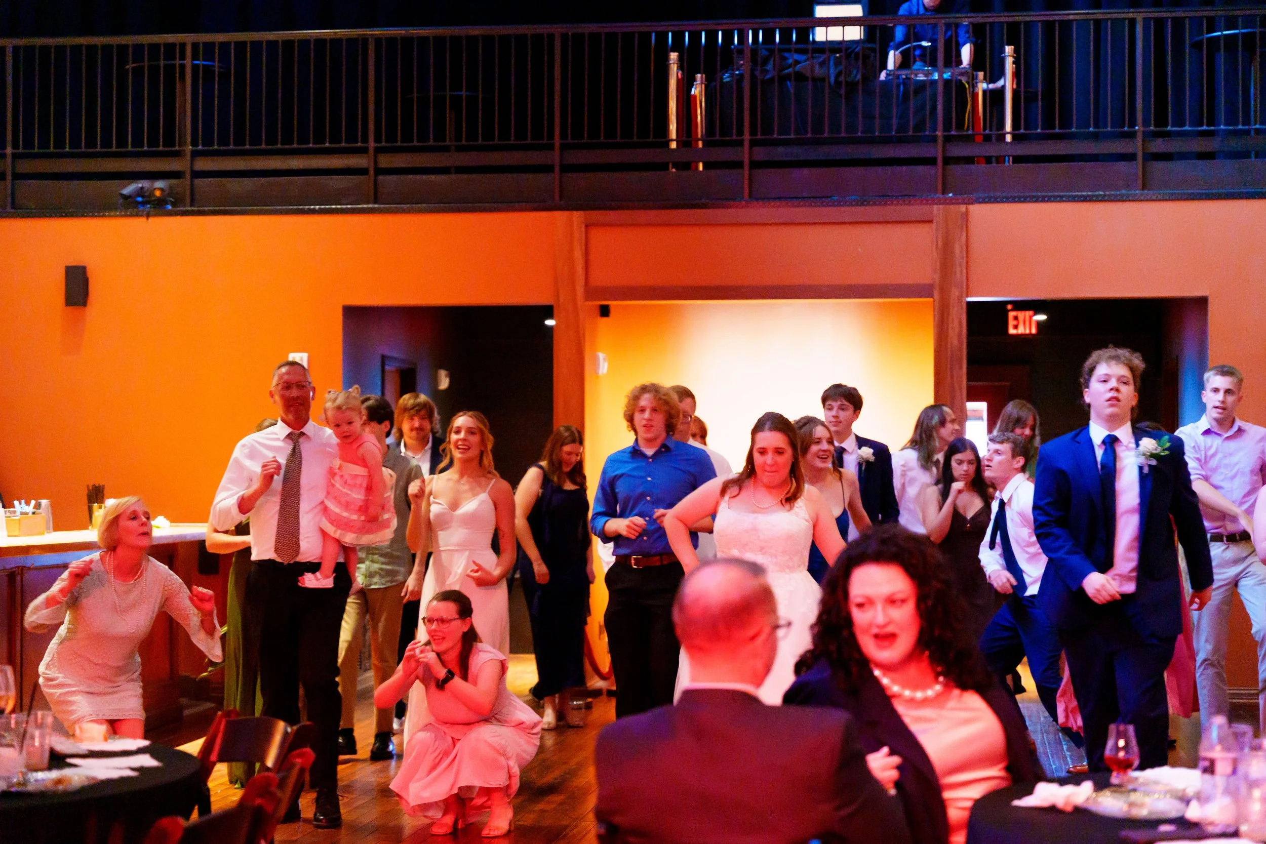 A group of people, including children and adults, are gathered at a party or celebration in a room with warm lighting. Some are smiling, dancing, or talking, with a person holding a young girl in the center. The room has orange and yellow walls, with