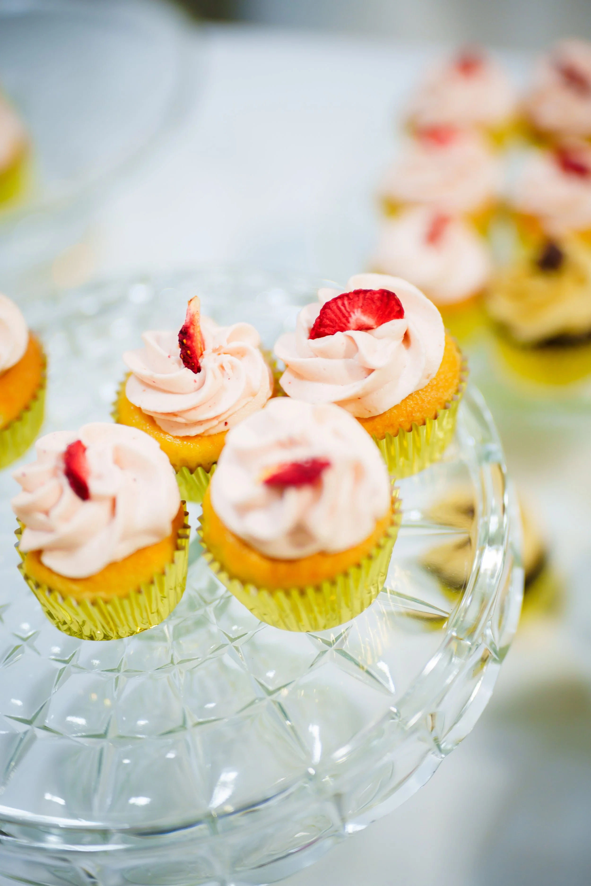Mini cupcakes with pink frosting topped with slices of strawberries on a glass cake stand.