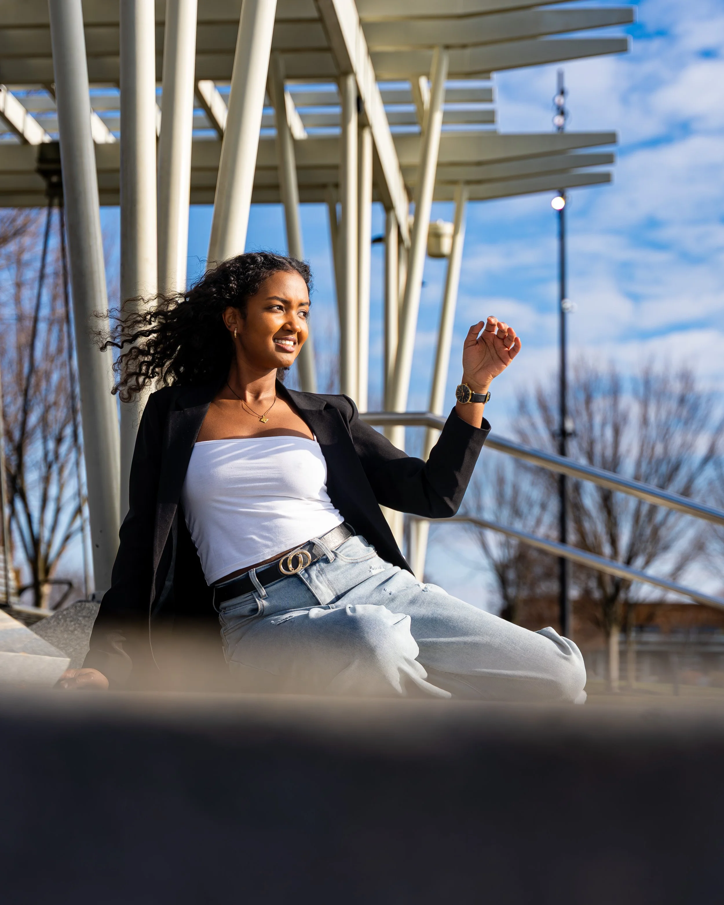 A young woman sitting on steps outdoors, smiling and looking to the side, with modern building architecture behind her and a clear blue sky.