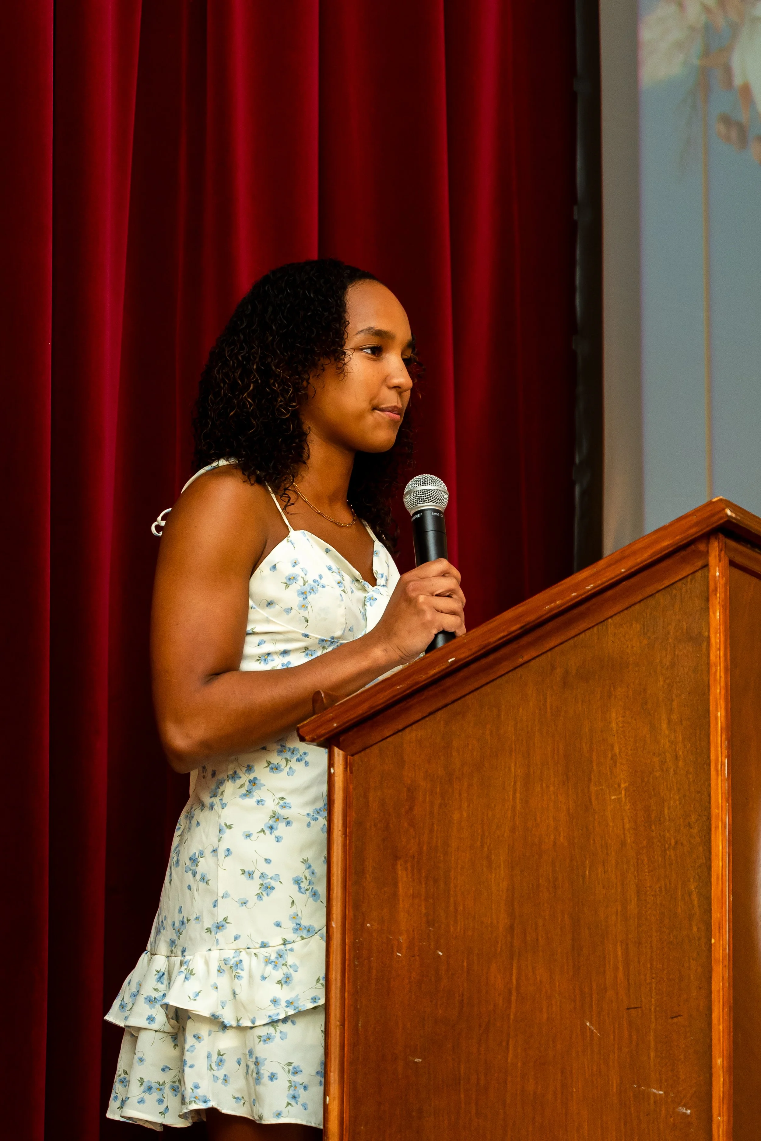 A young woman with dark, curly hair stands at a wooden podium, holding a microphone, in front of red stage curtains, wearing a white dress with blue floral patterns.
