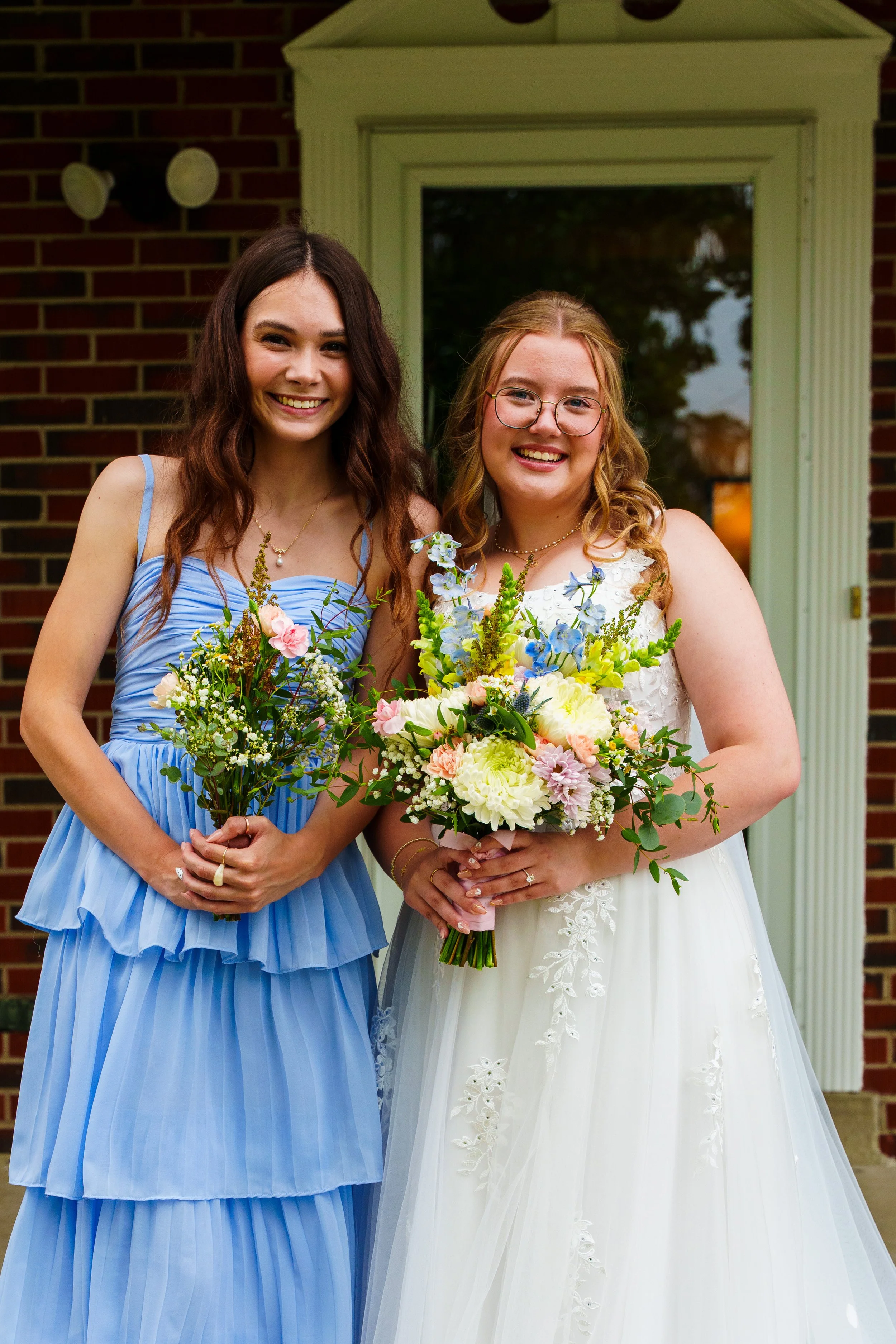 Two women in formal dresses holding bouquets of flowers, standing outside in front of a brick building, smiling at the camera.