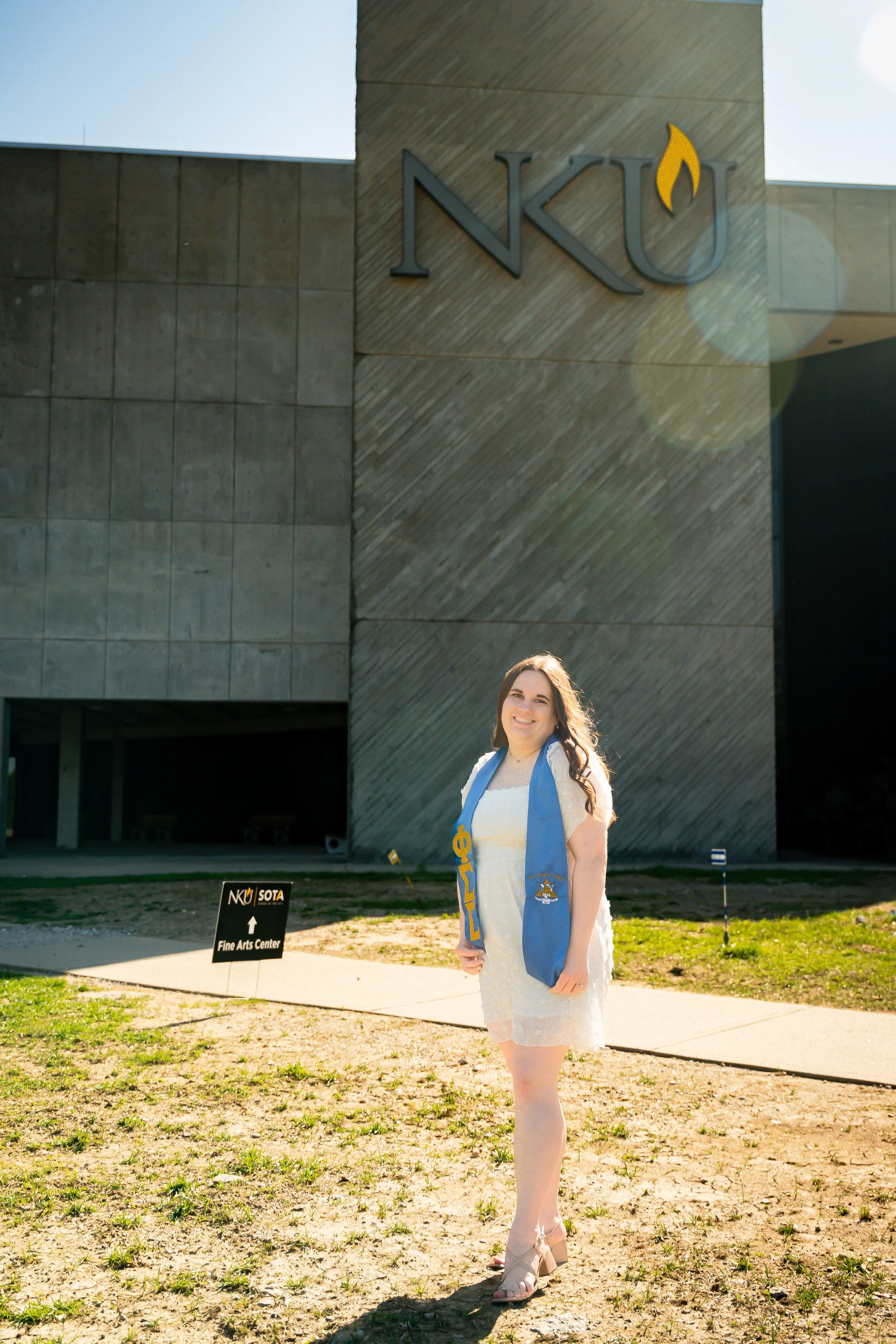 A young woman with long brown hair wearing a white dress and beige heels, standing outside in front of a modern building with a sign that reads 'NKU' and 'Fine Arts Center.' The woman has a blue and yellow scarf around her neck with a university embl