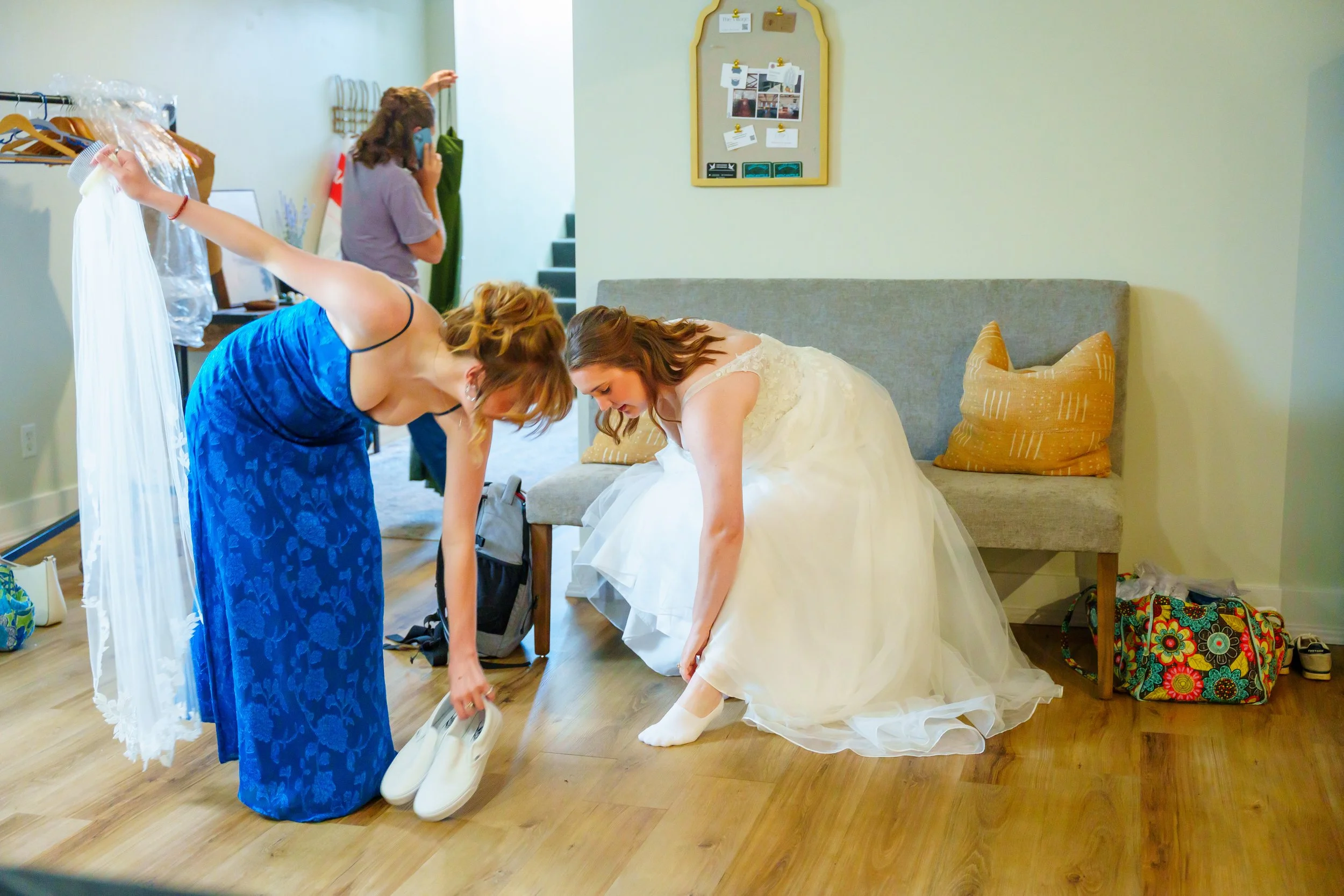 A bride in a white wedding dress is crouching and putting on white shoes in a room, while a woman in a blue dress is assisting her. Another person in casual clothing is talking on the phone in the background. The room has a gray sofa with orange pill