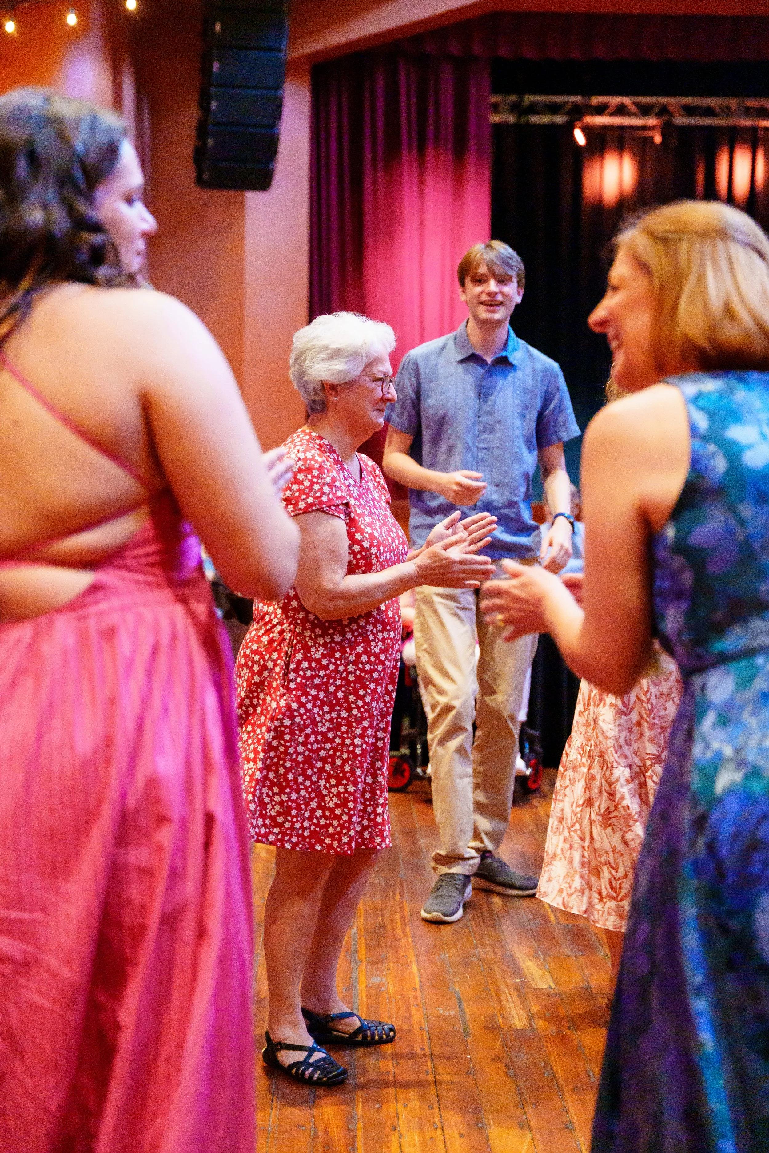 Older woman in red and white dress talking to younger women in colorful dresses, with a young man in a blue shirt smiling in the background, inside a hall with stage curtains.