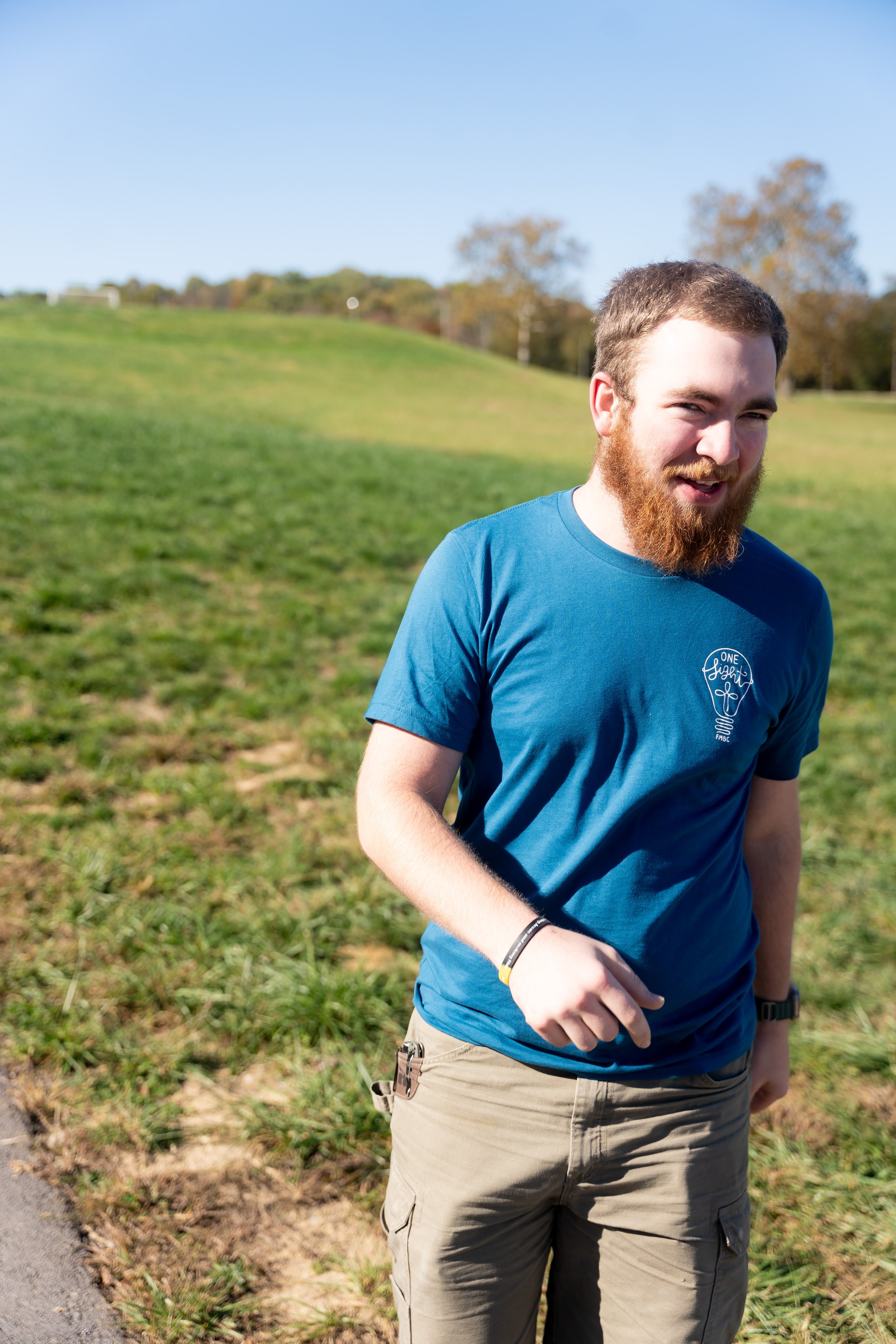 A young man with a beard walking outdoors on a sunny day, wearing a blue t-shirt and beige cargo shorts, with trees in the background and grass on the ground.