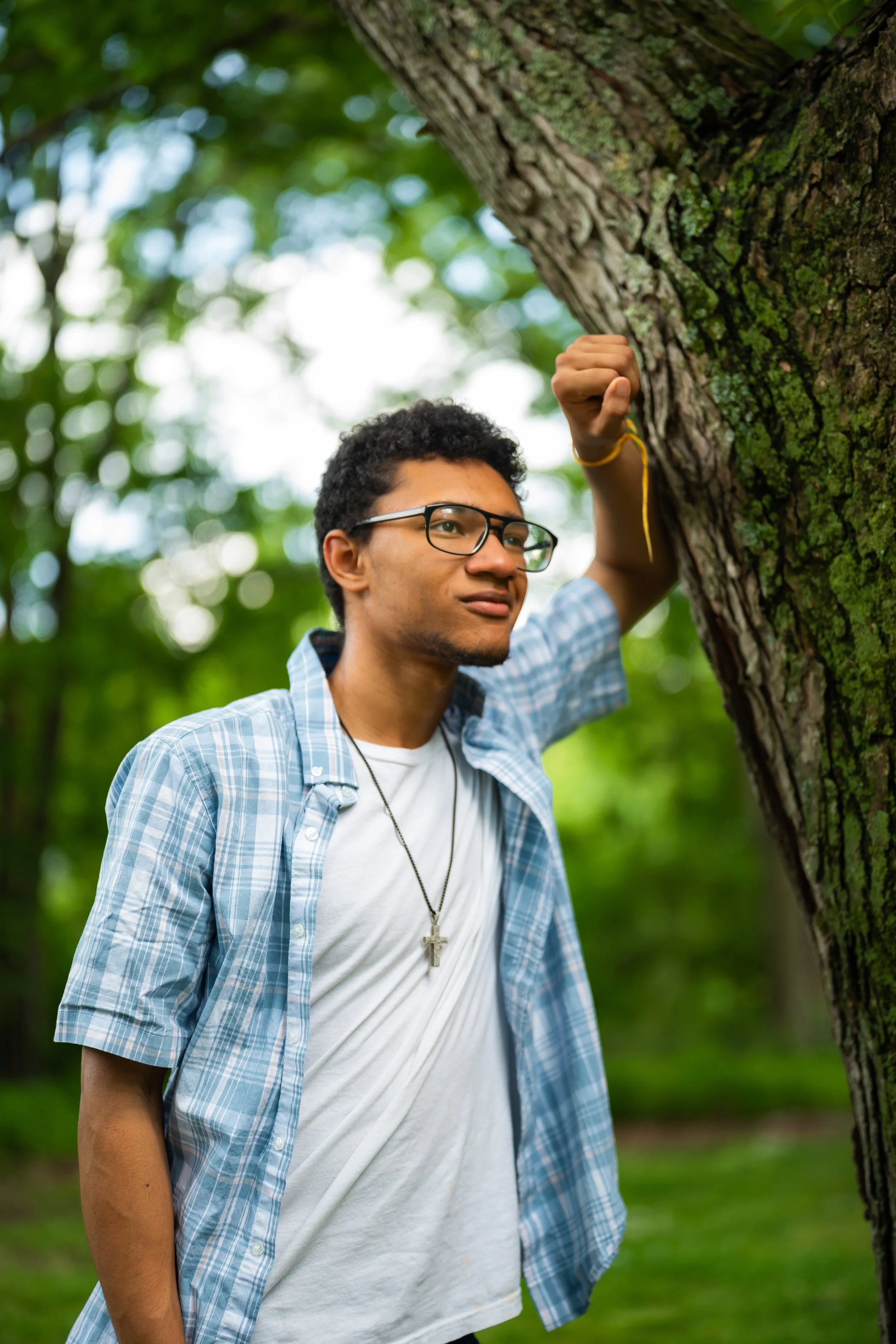 A young man with curly black hair, glasses, and a cross necklace, wearing a white t-shirt and a light blue plaid shirt, is outdoors in a wooded area, touching or holding onto a tree trunk with moss and rough bark.