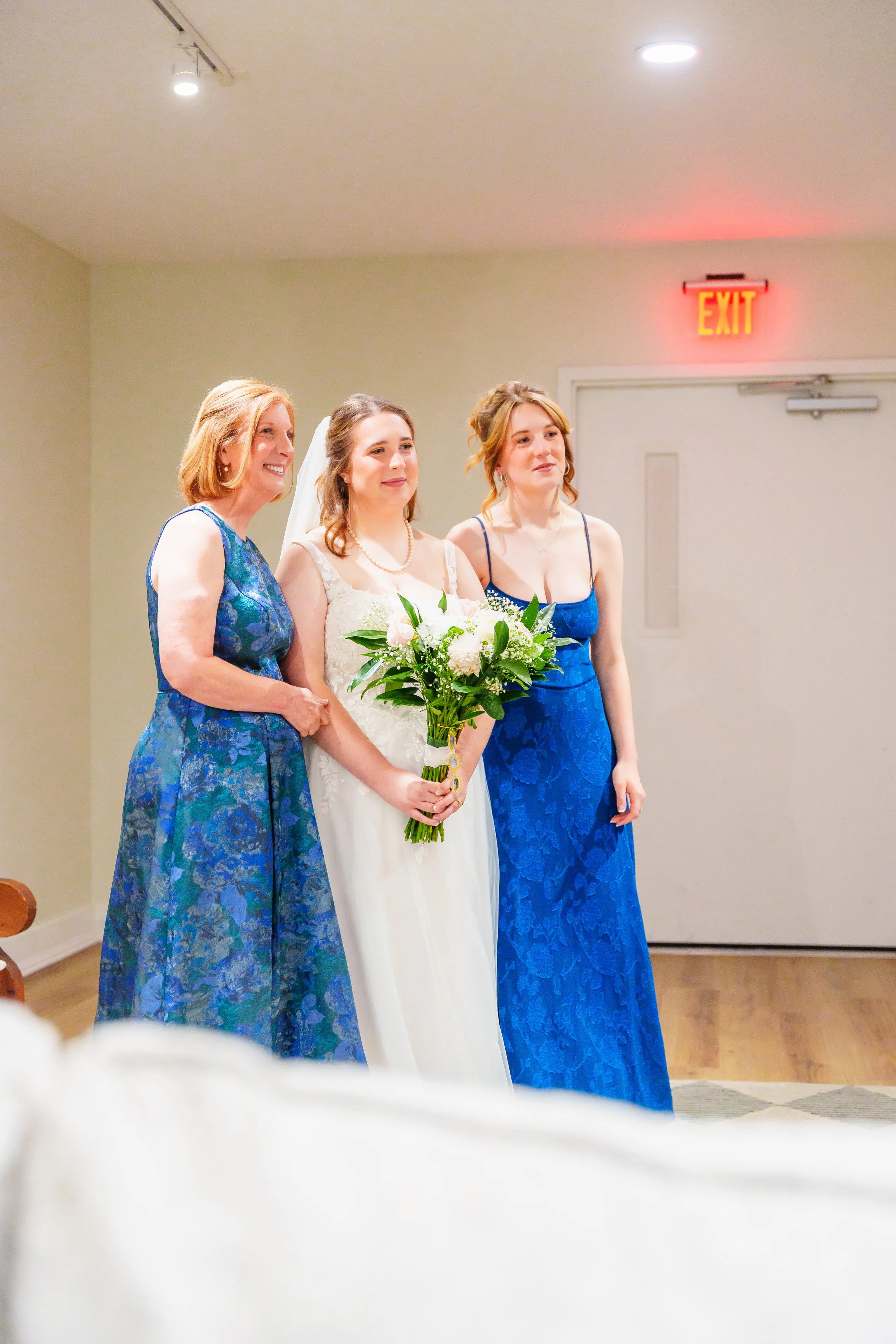 Three women, one in a wedding dress holding a bouquet, standing next to two women in blue dresses inside a room with a beige wall and an exit sign above a door.