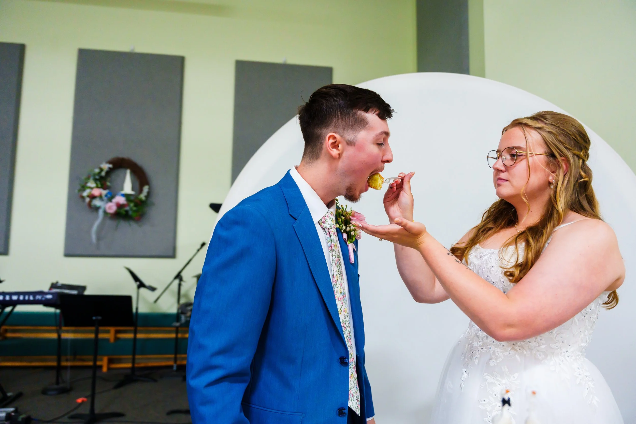 A bride with glasses, wearing a white lace wedding dress, feeding a cake or dessert to a groom in a blue suit with a floral tie, during their wedding ceremony.