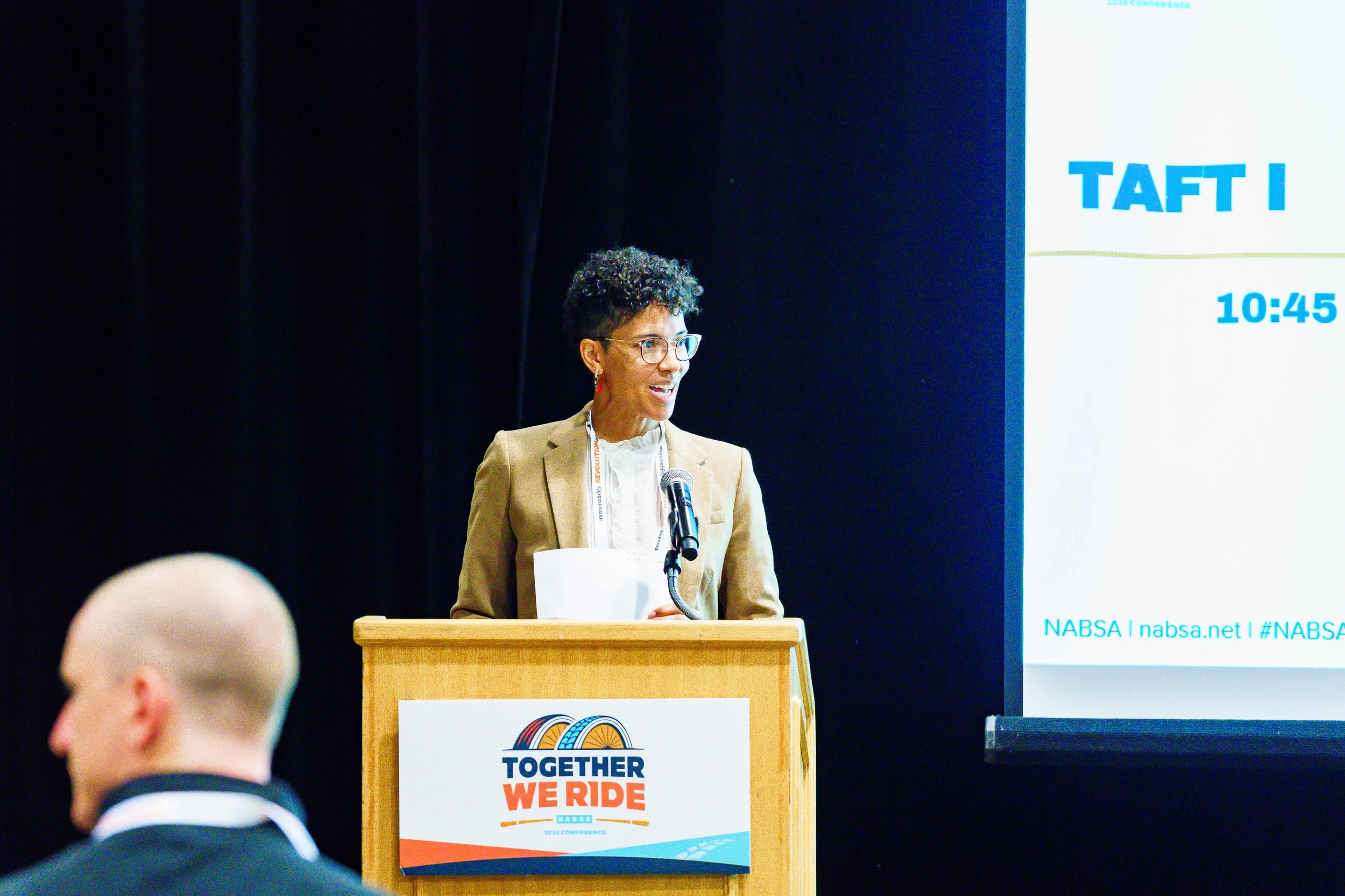 A woman with short curly hair and glasses speaking at a podium during a conference. Behind her is a large screen displaying the text 'NABSA | nabsa.net | #NABSA'. The podium has a sign with the logo and the slogan 'Together We Ride.' She is wearing a tan blazer and red earrings.