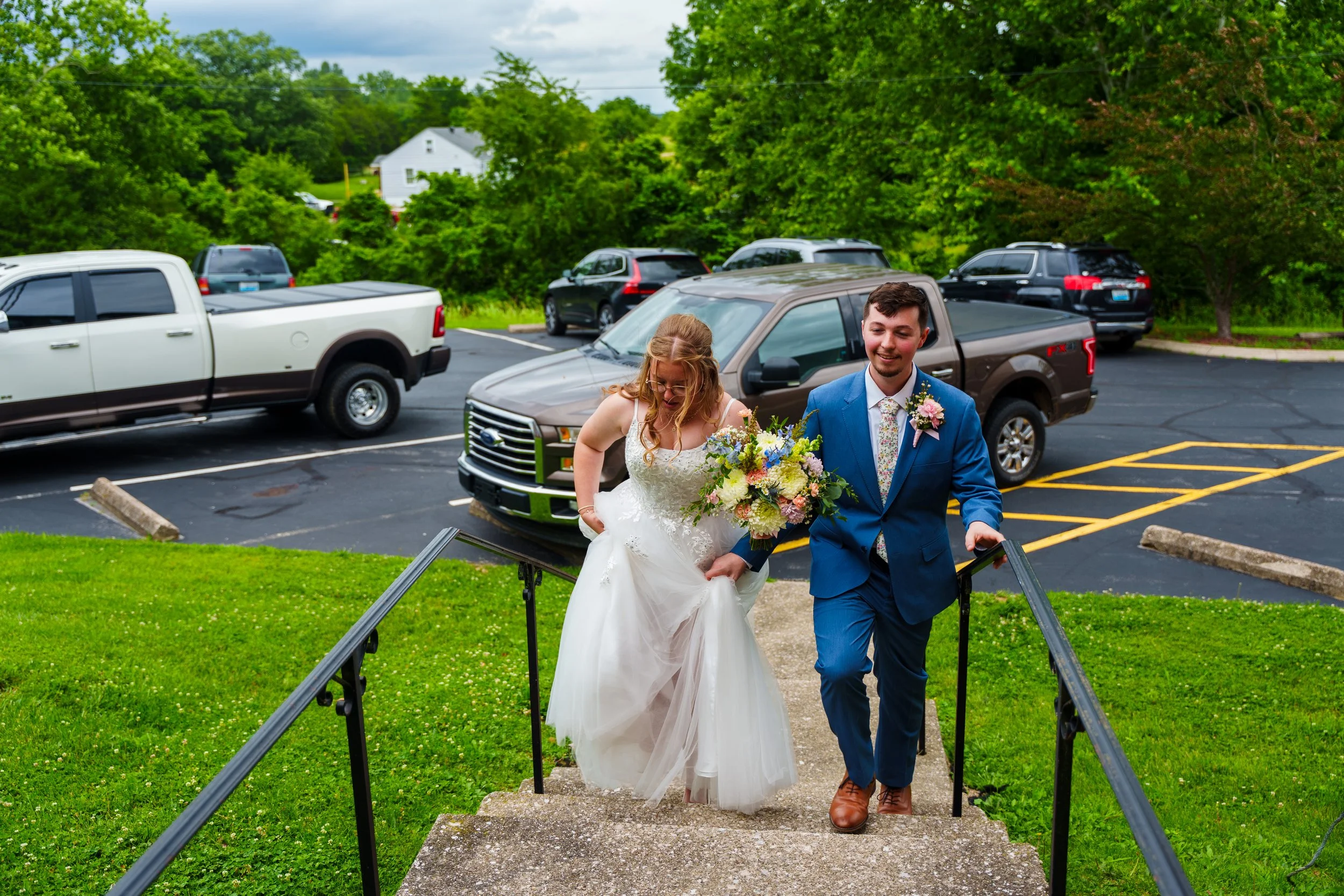 A newlywed couple walking up a small set of outdoor stairs, the bride in a white wedding dress holding a bouquet and the groom in a blue suit with a floral tie. They are smiling and surrounded by a parking lot with several cars and lush green trees i