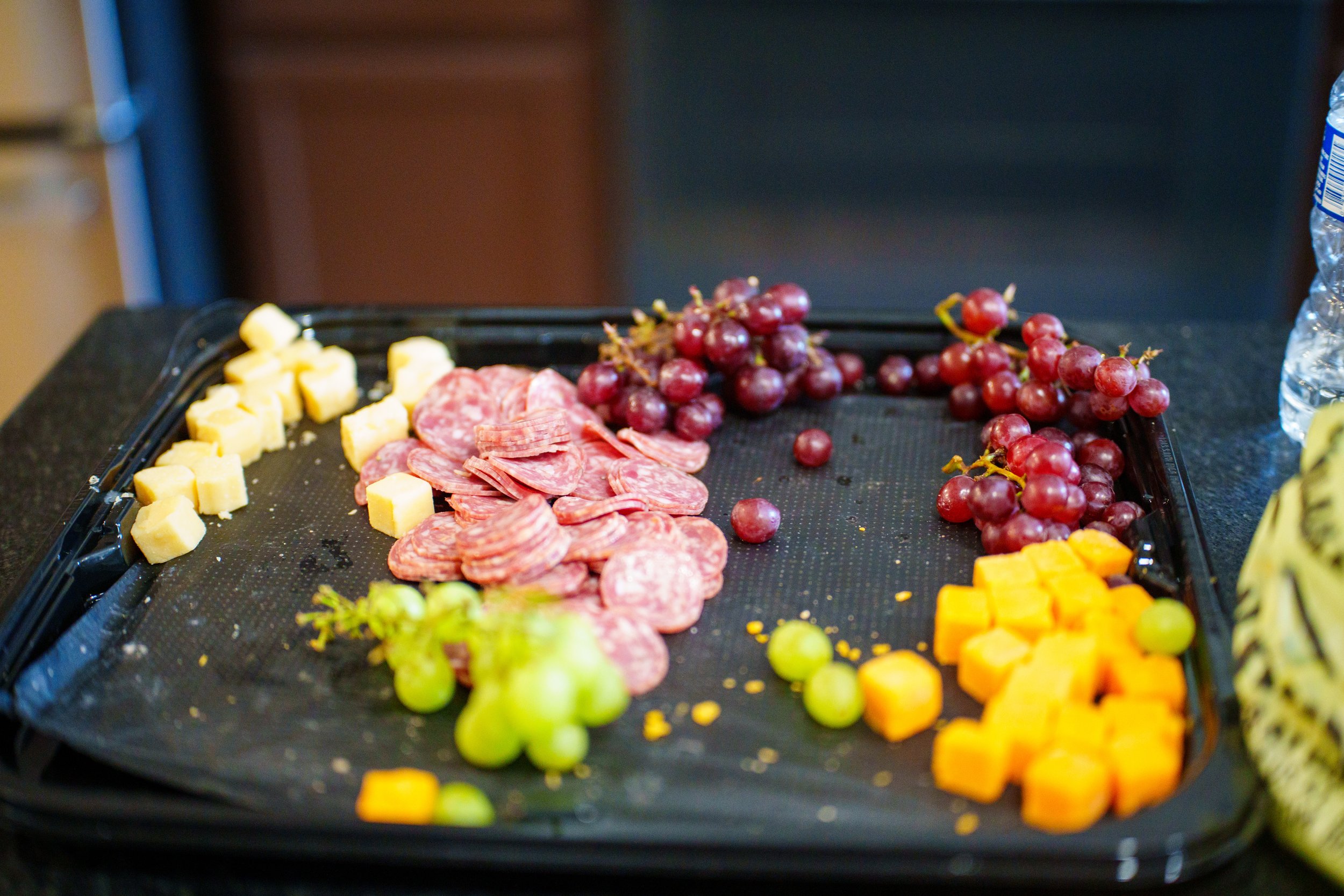 A black serving tray with cubes of cheese, slices of salami, and bunches of red and green grapes.