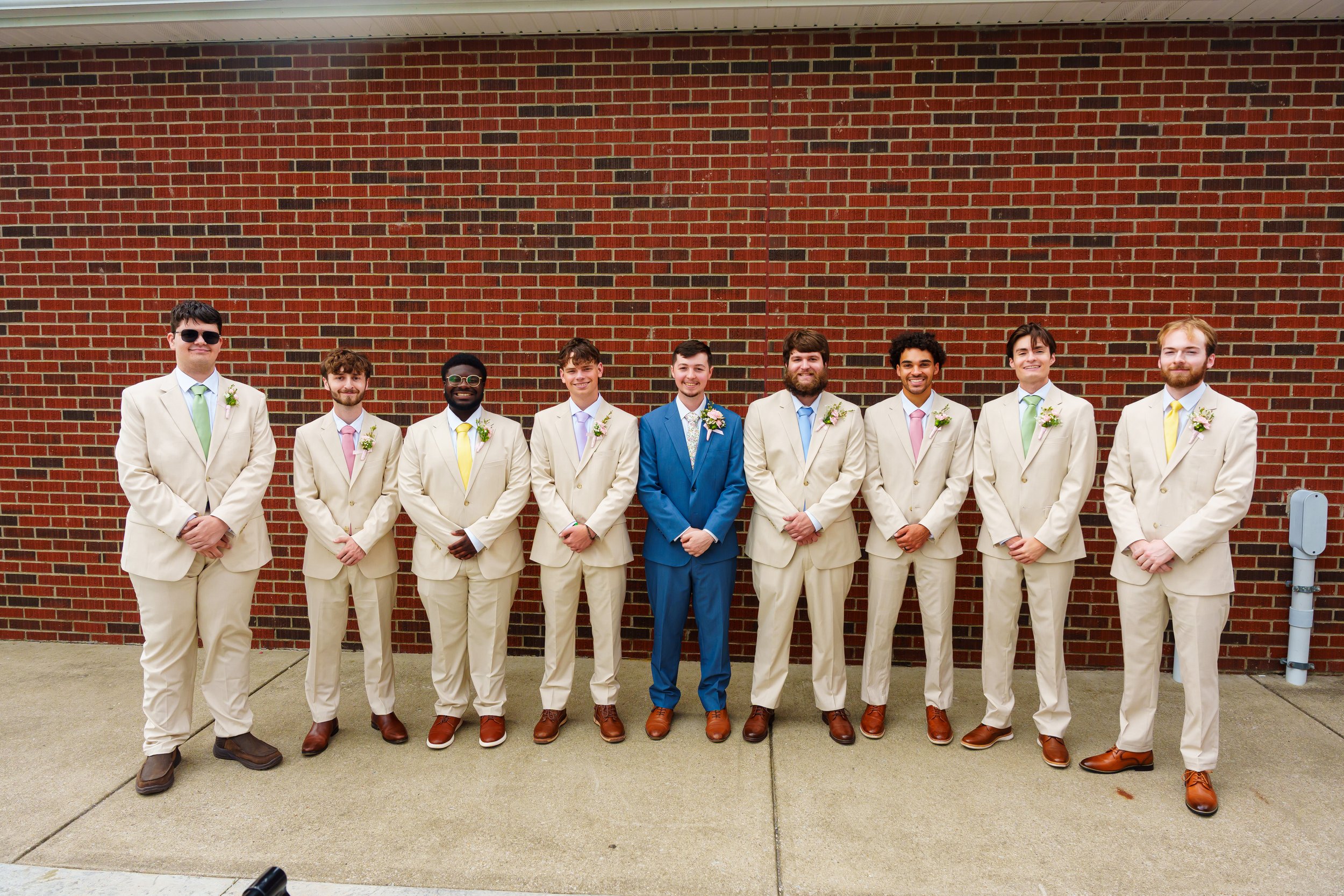A group of nine men dressed in formal suits, standing in front of a brick wall, likely at a wedding or special event.