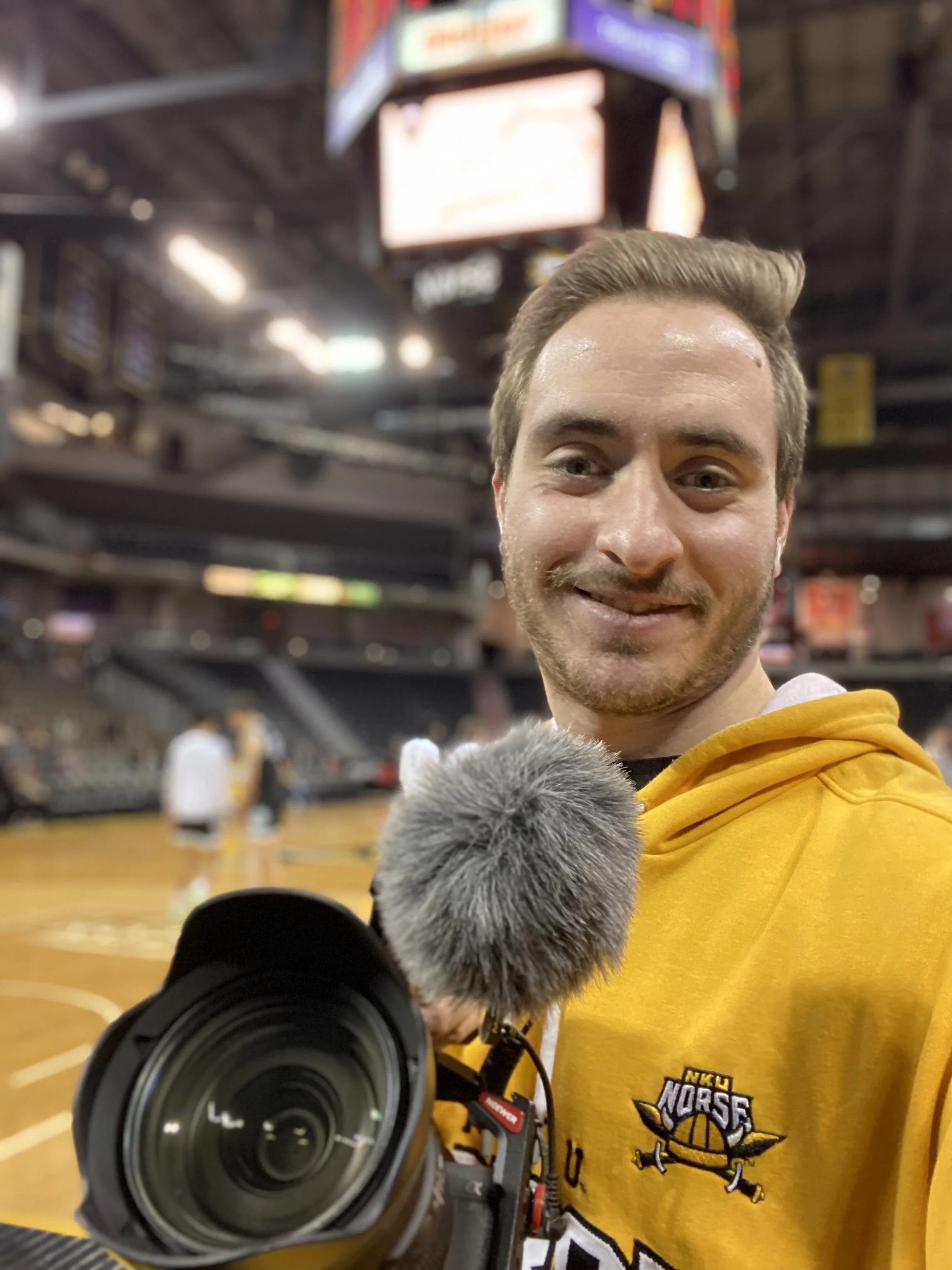 A man with light brown hair and a short beard smiling, taking a selfie with a camera in an indoor sports arena. He is wearing a yellow hoodie with the NKU Norse logo. The background shows bleachers, electronic scoreboards, and other people.