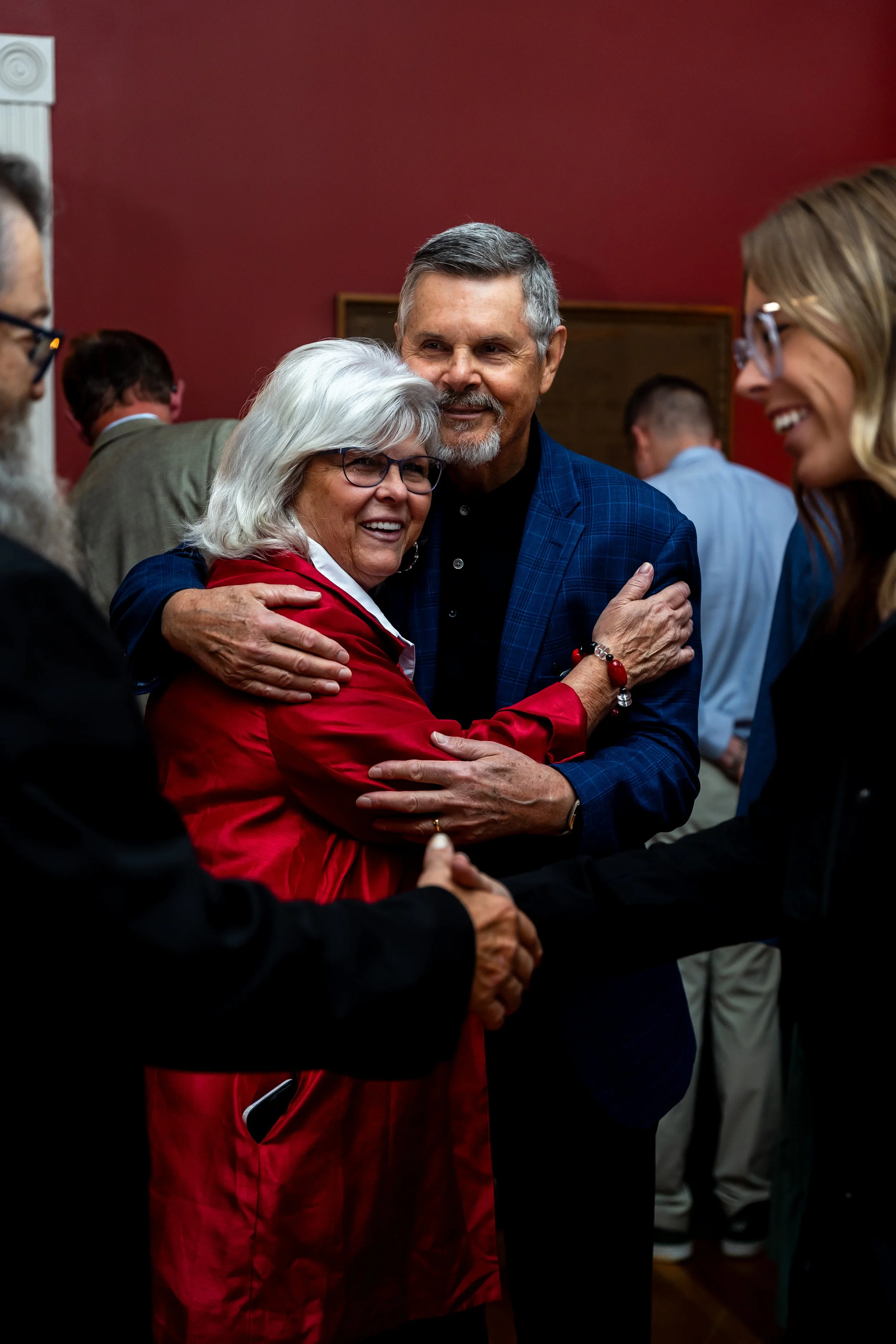 People engaging in a warm hug or conversation at a social gathering, with a focus on an elderly woman in a red jacket and a middle-aged man in a blue checked blazer.
