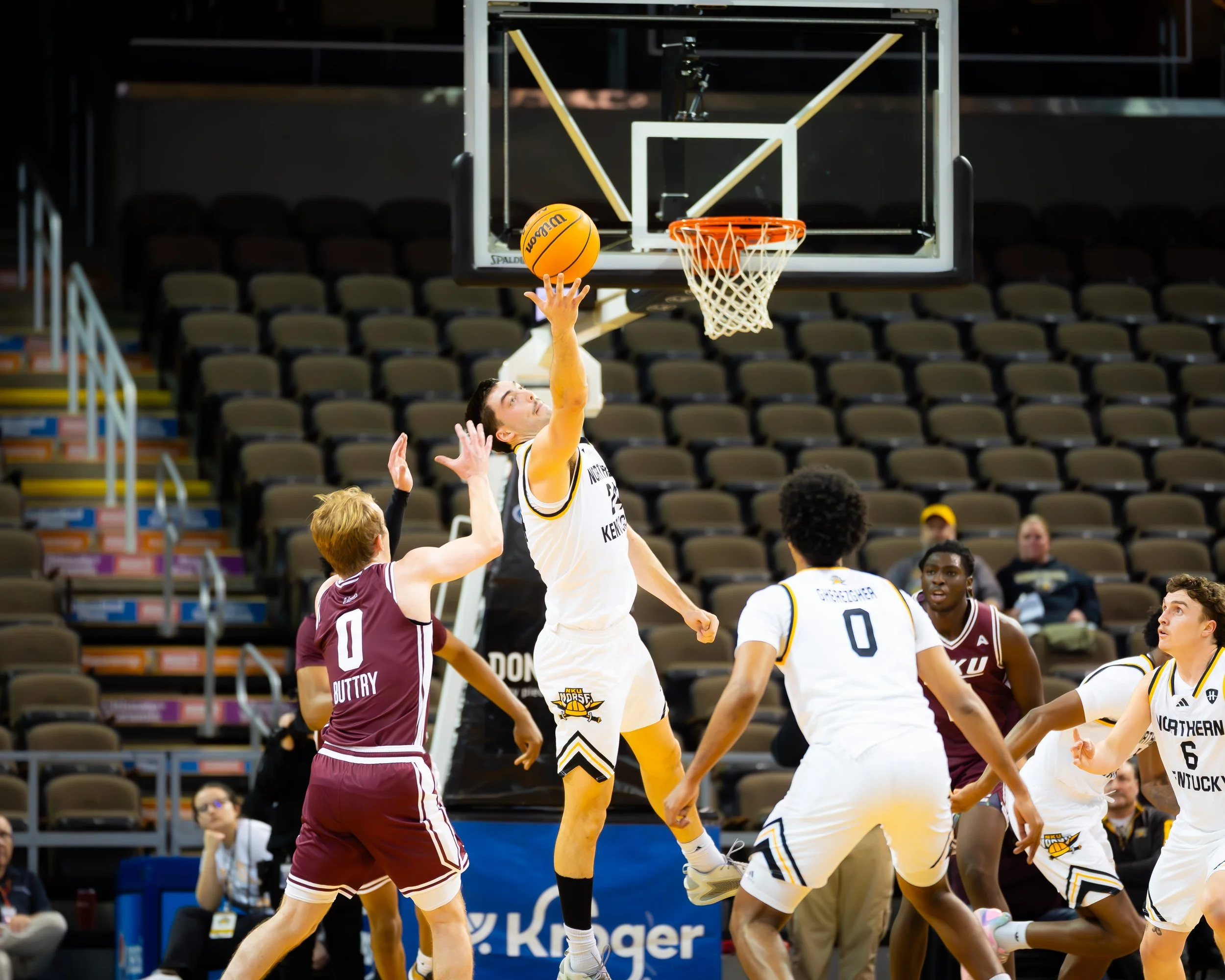 A basketball game with players in white and maroon jerseys competing near the hoop, with one player in white jumping to shoot or dunk while others attempt to block or defend.