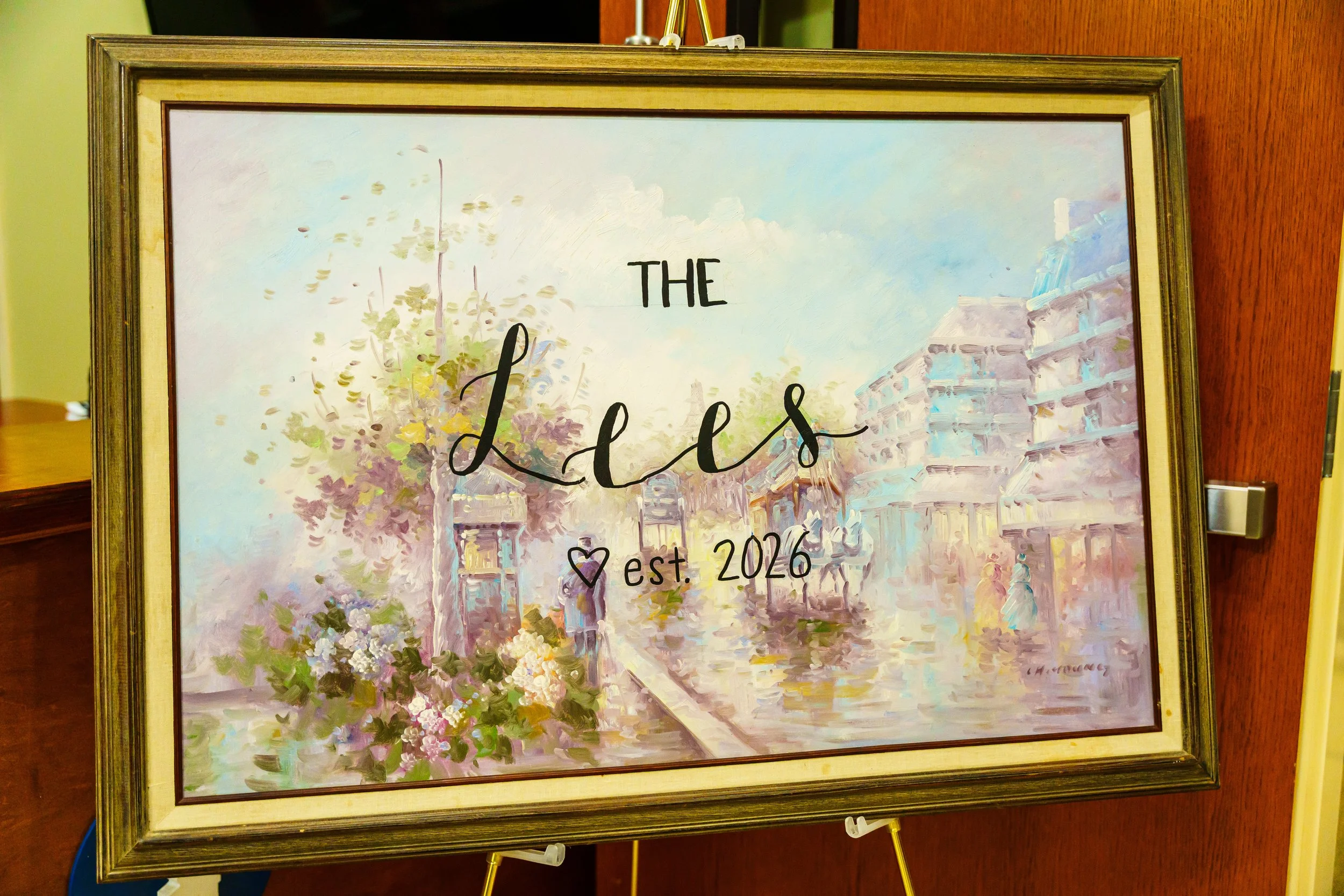 A framed painting of a street scene with buildings, trees, and flowers, featuring the text 'The Ladies' and 'est. 2026' in black, inside a gold-colored frame, leaning on a stand against a wooden-paneled wall.