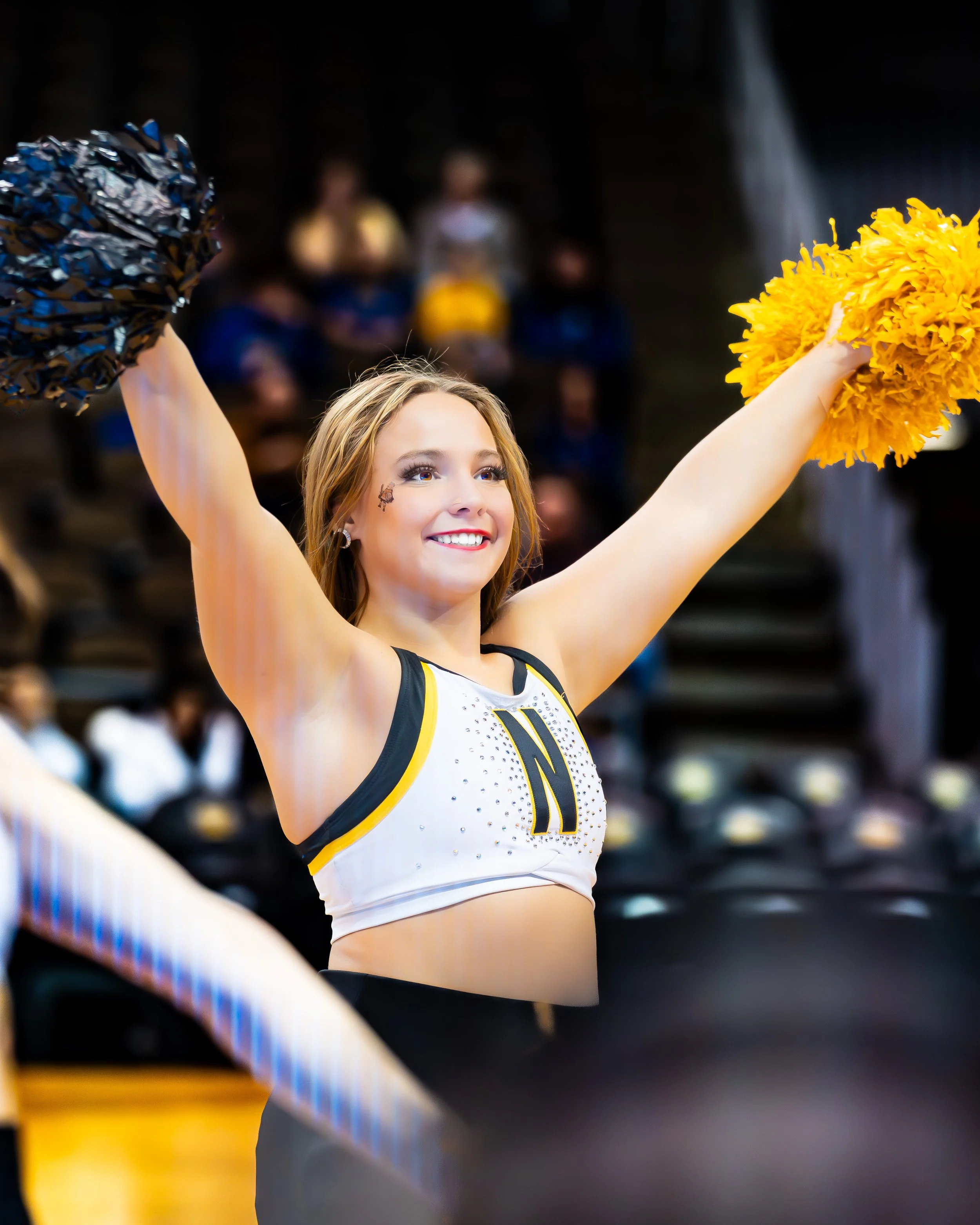 A cheerleader in a black and white uniform with a yellow 'N' on the front, smiling and holding pom-poms, celebrating at a sports event.
