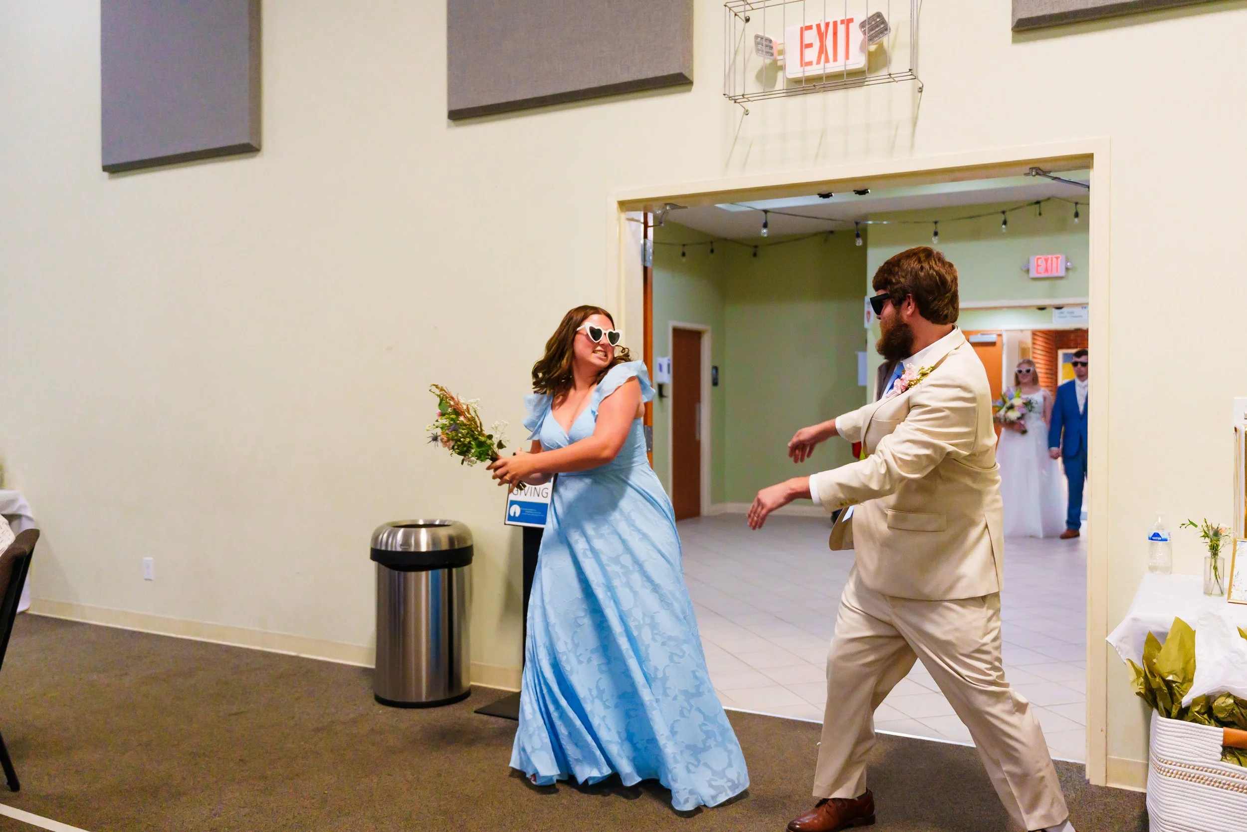 A woman in a light blue dress with heart-shaped sunglasses holding a bouquet, dancing with a man in a beige suit and sunglasses at a wedding reception.