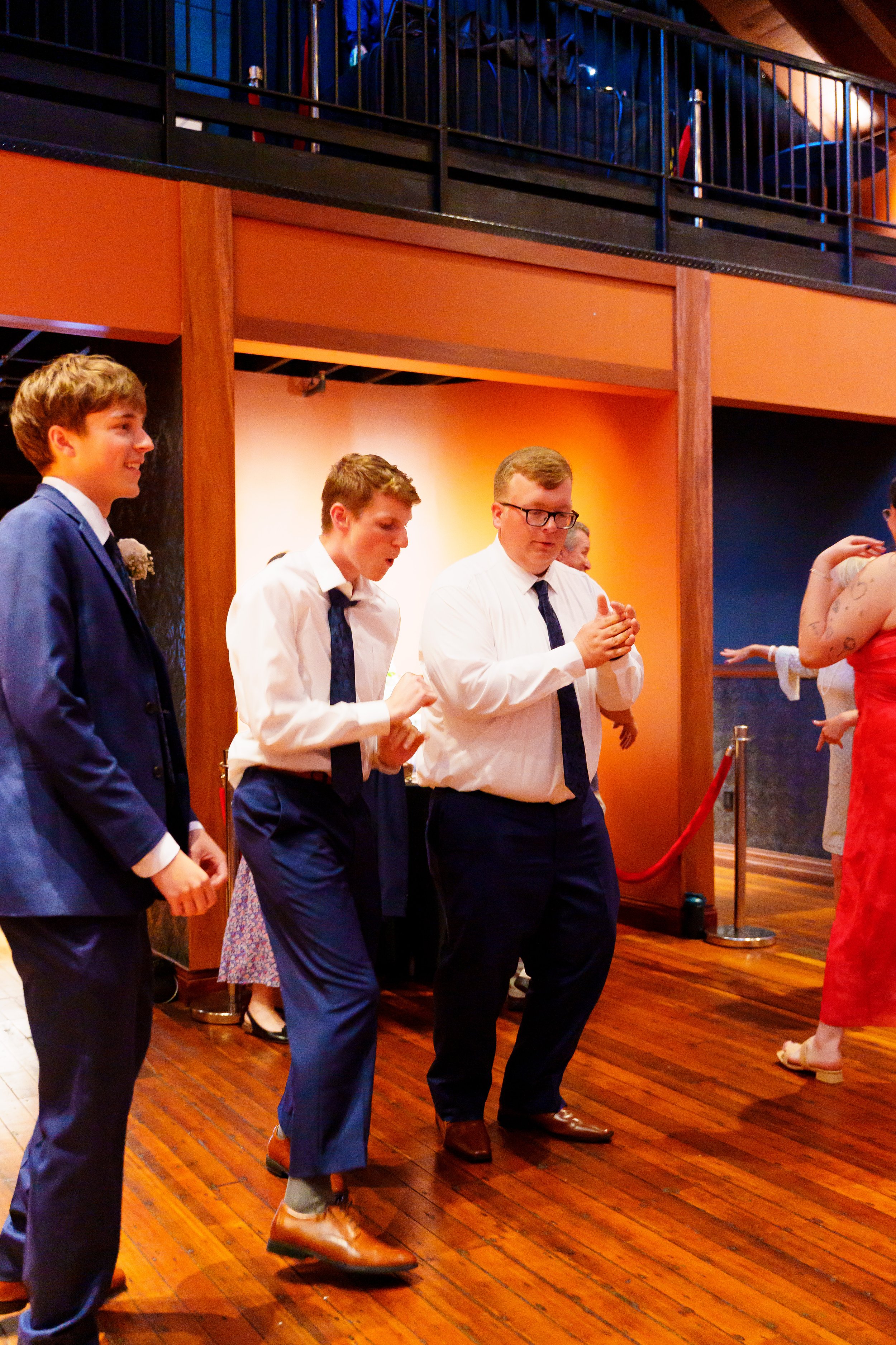 A group of young men dressed in formal attire, standing on a wooden floor at an event. They are talking and appear to be enjoying themselves. The background features a warm-colored wall and an upper balcony with metal railings.