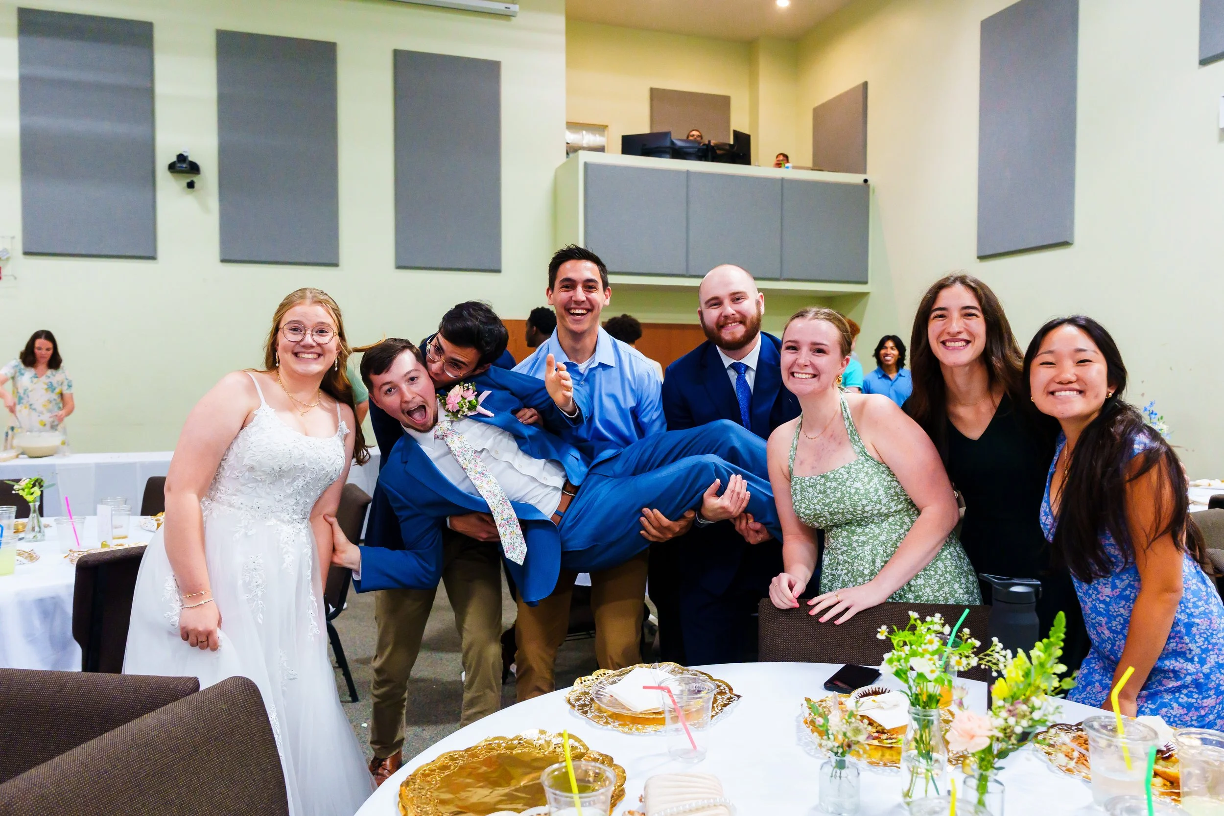 Group of eight smiling friends at a celebration, with one person being lifted, in a decorated hall with tables of food and drinks.