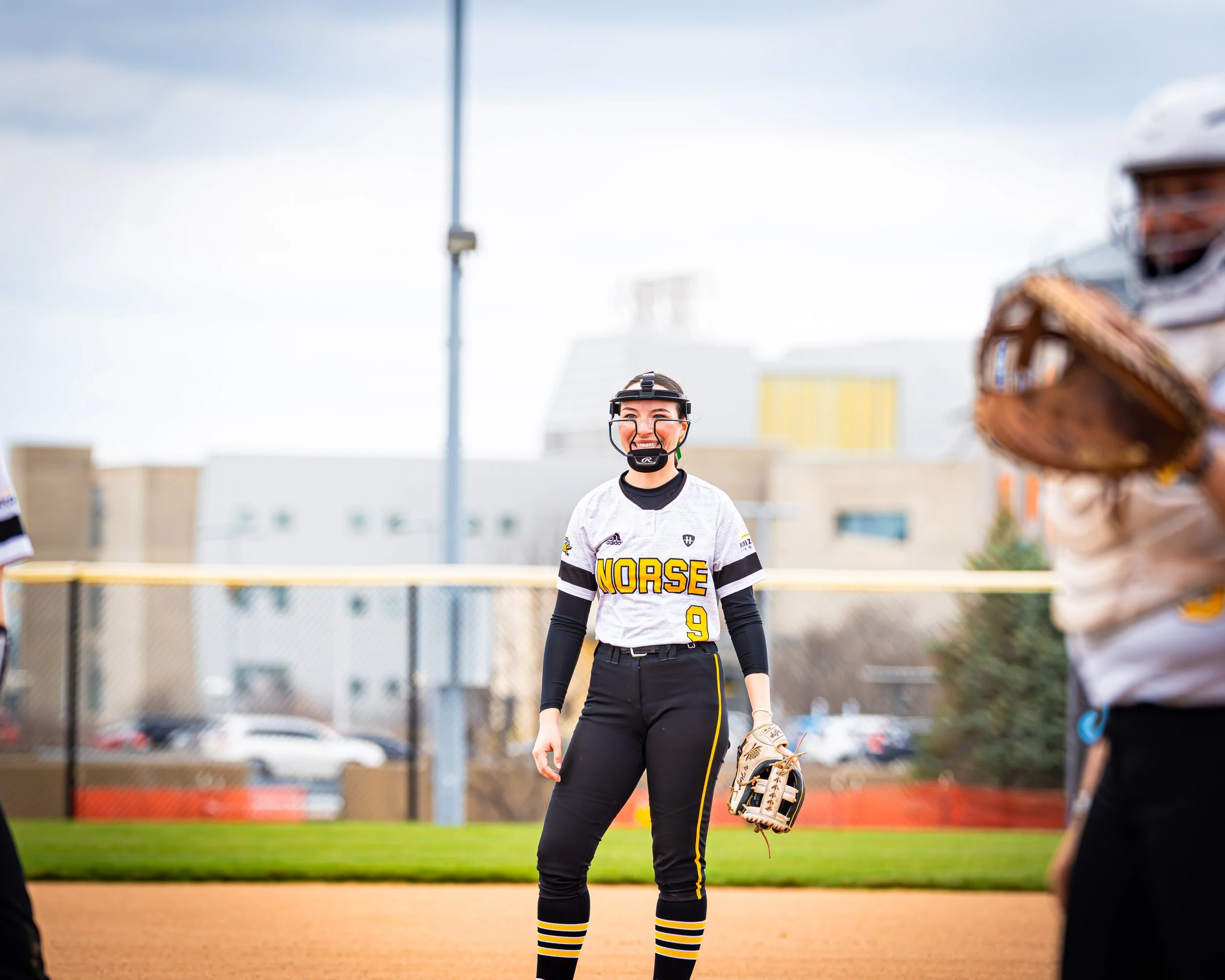 A female softball player standing on the field with a smile, wearing a white jersey with the word "NORSE" and the number 9, black pants with yellow stripes, and striped socks. She's holding a softball glove and a baseball cap, with other players and 