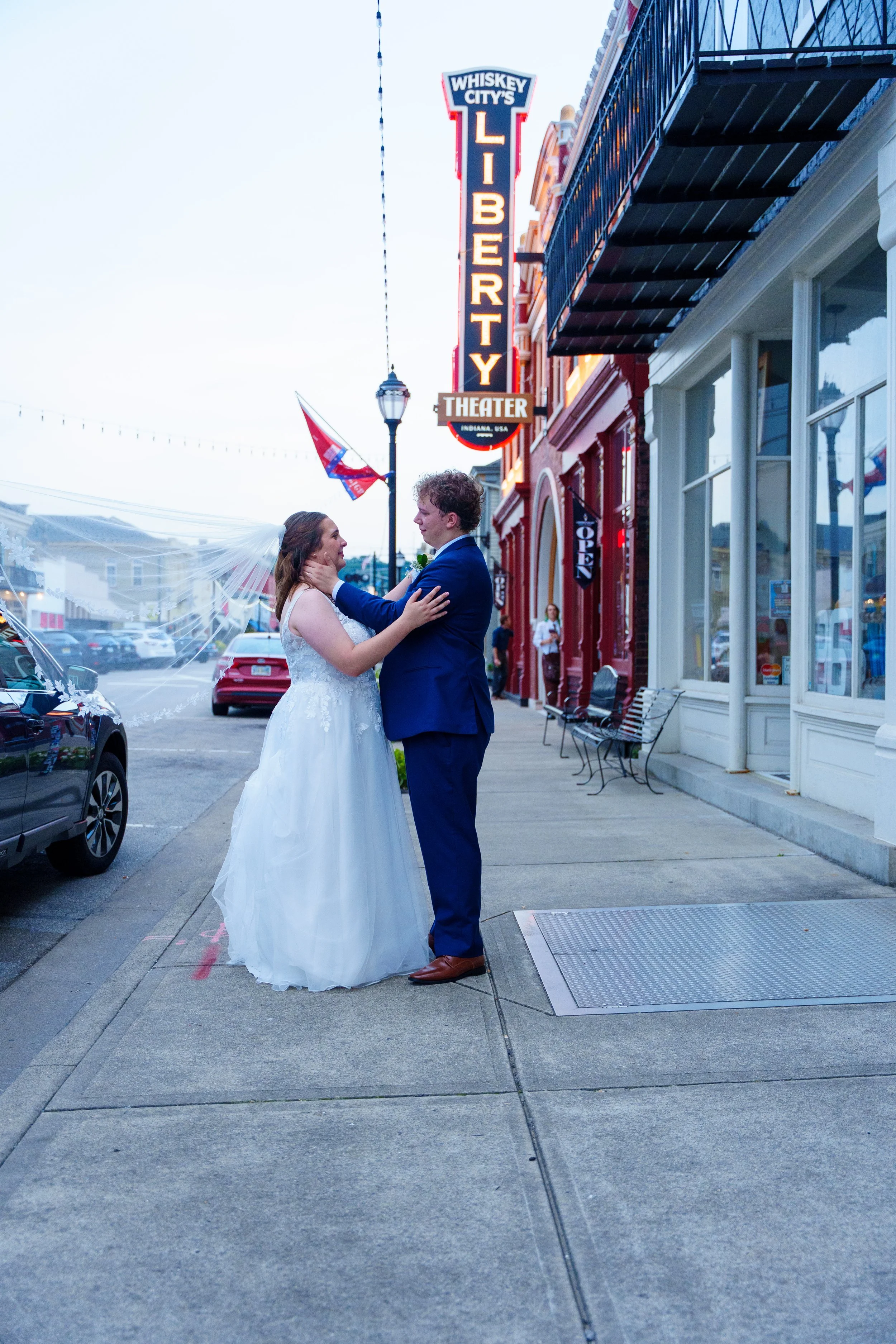 A bride and groom share a moment on a city sidewalk in front of a theater with a sign that reads "Whiskey City's Library." The bride wears a white wedding dress with a veil, and the groom is dressed in a blue suit. They are smiling and looking into e